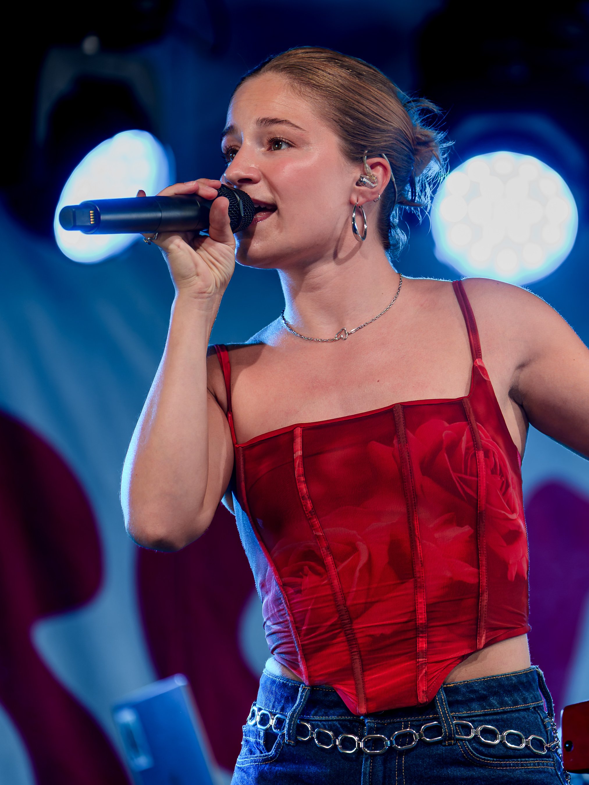 En ung kvinde synger i en mikrofon med lyst hår bundet tilbage, iført en rød top og blå jeans med kæde i taljen, under koncert med blå baggrundsbelysning.