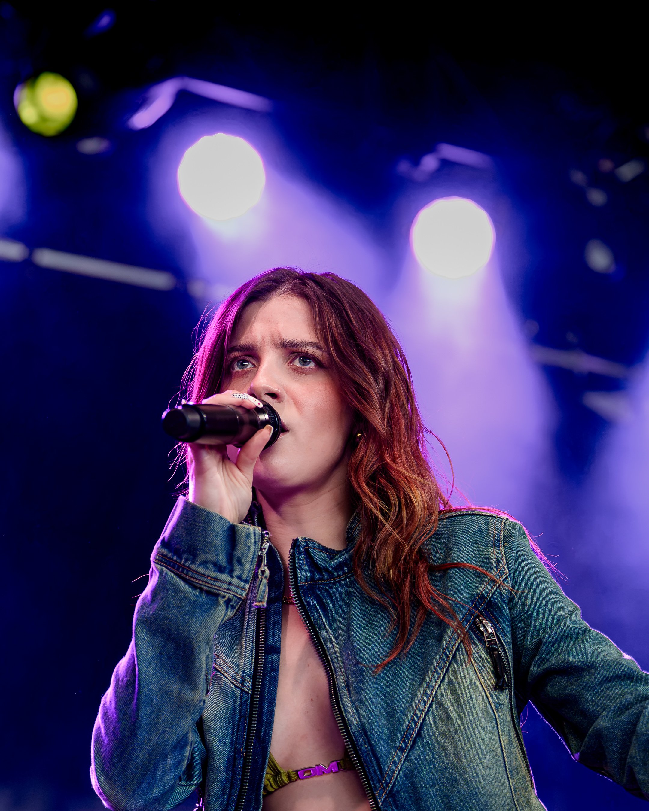 En ung kvinde med rødt hår, iført denimjakke, holder en mikrofon og ser op mod scenen, foran farvede stage-lys.