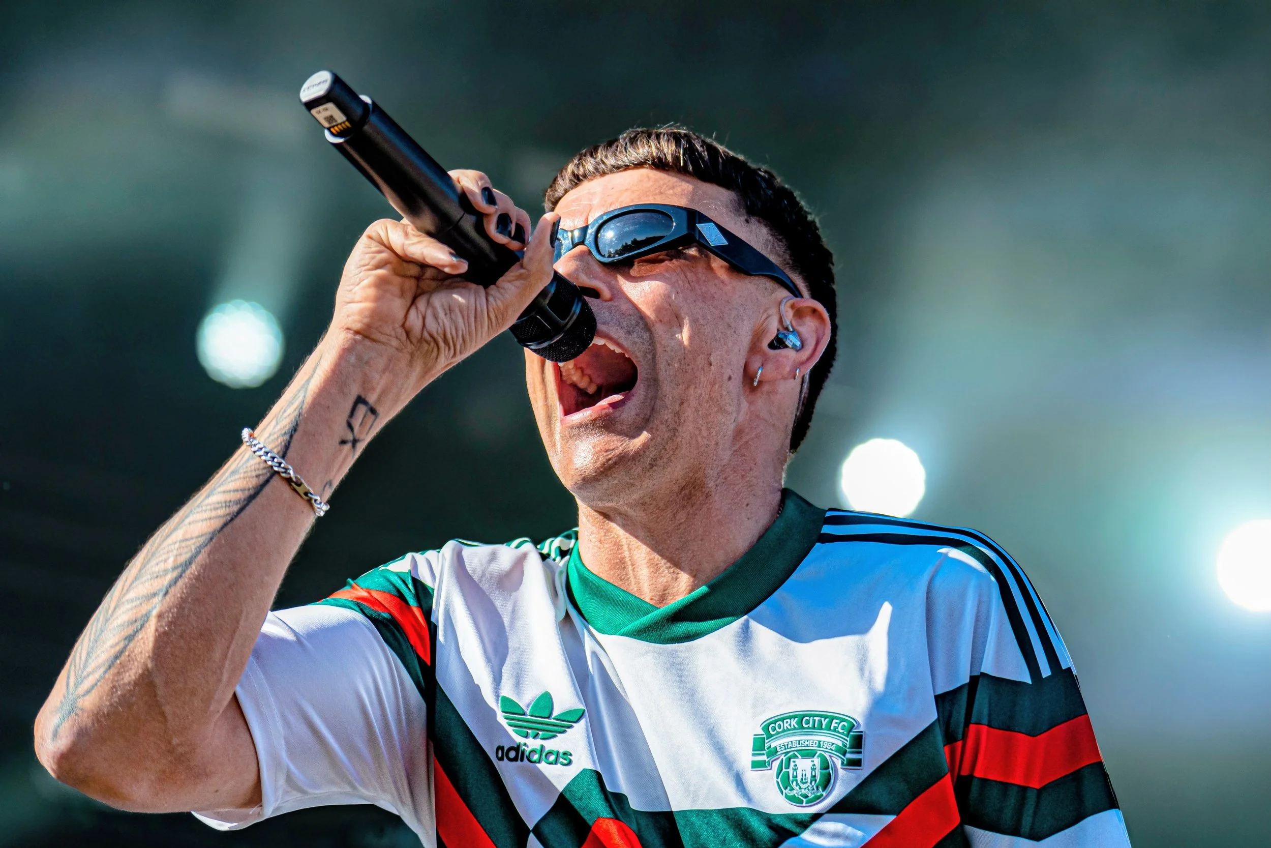 En person i en hvid, grøn, rød og sort trøje med Cork City FC-logo og Adidas-logo tager et mundfuld af en mikrofon, ved sol- eller stadionlys.