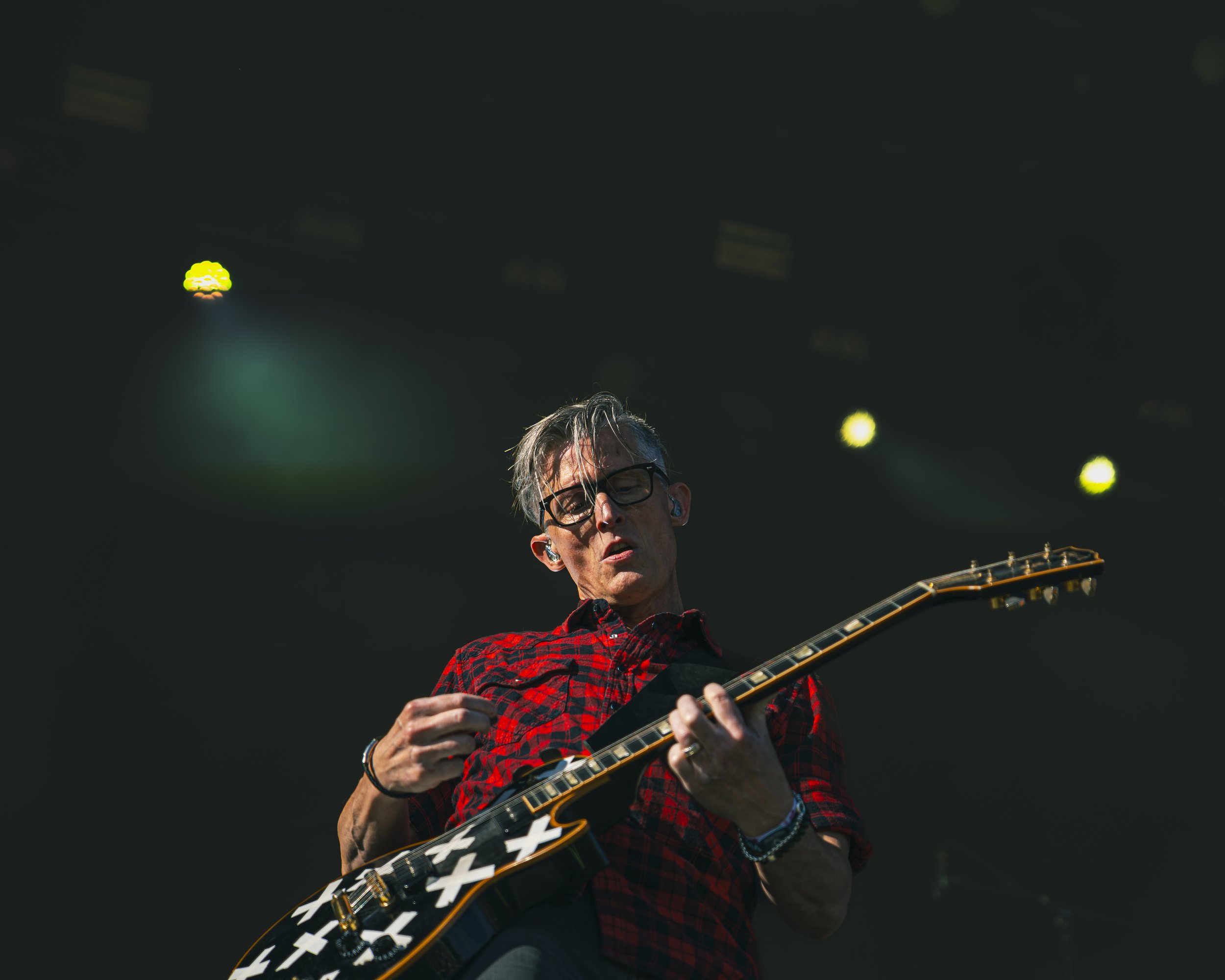 Person spiller guitar på en koncertscene, iført rød og sort ternet skjorte, og har briller og lyst hår.