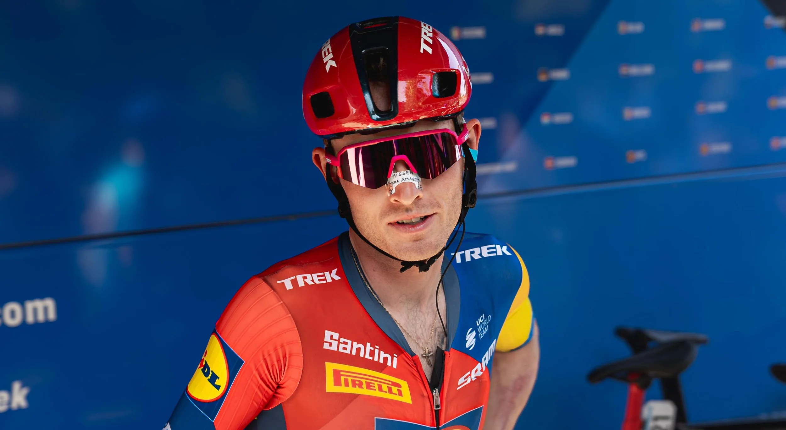 Cyklist i professionelt racing udstyr med hjelm, goggles og en jersey med sponsorlogoer, står foran en blå baggrund.