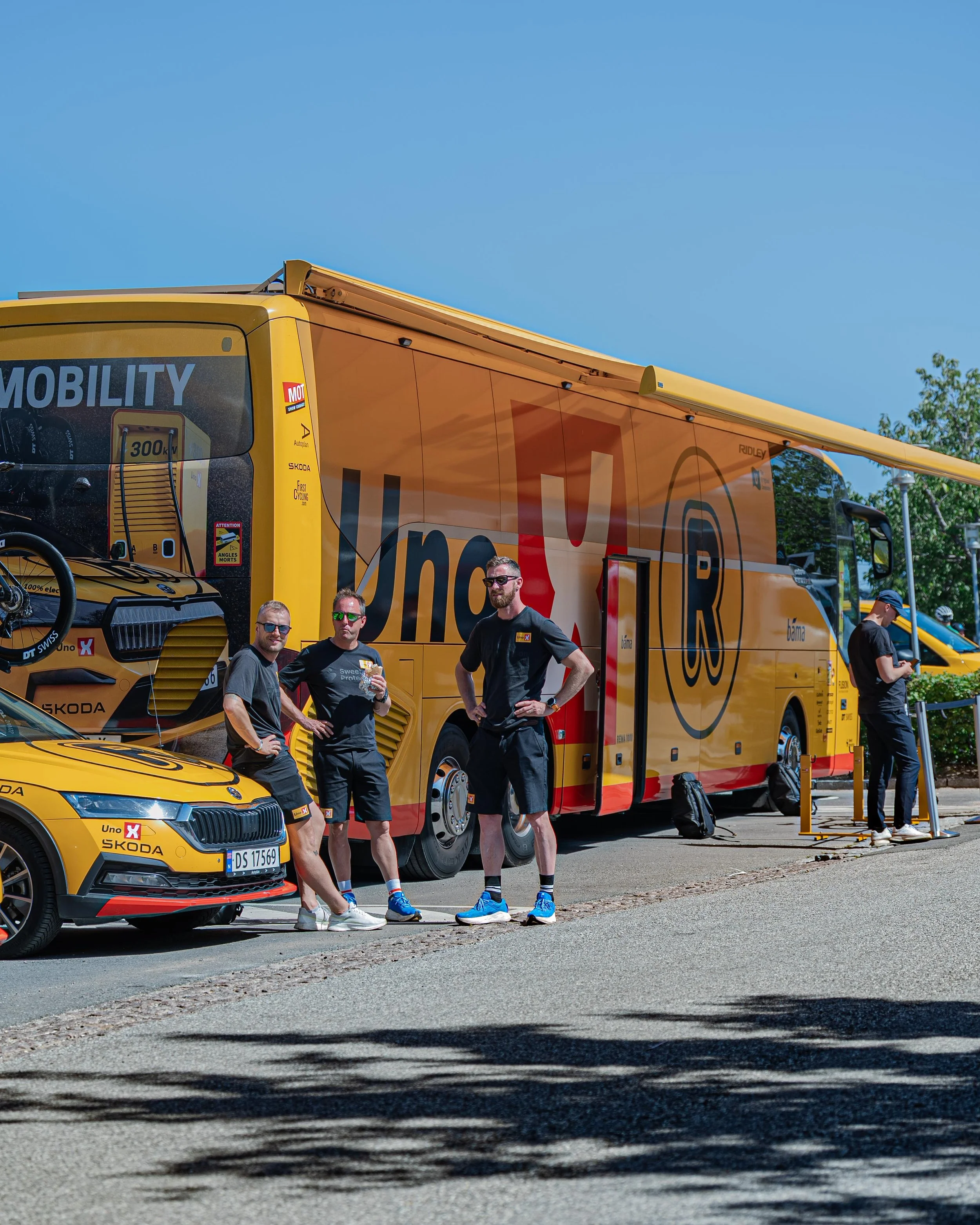 En gruppe mennesker står foran en større, gul Loeb-bus med branding for et professionelt hold. De er iført sorte T-shirts og solbriller, nogle holder drikkevarer, mens andre er ved at possessere bagage. Baggrunden viser klar blå himmel og grønne træe
