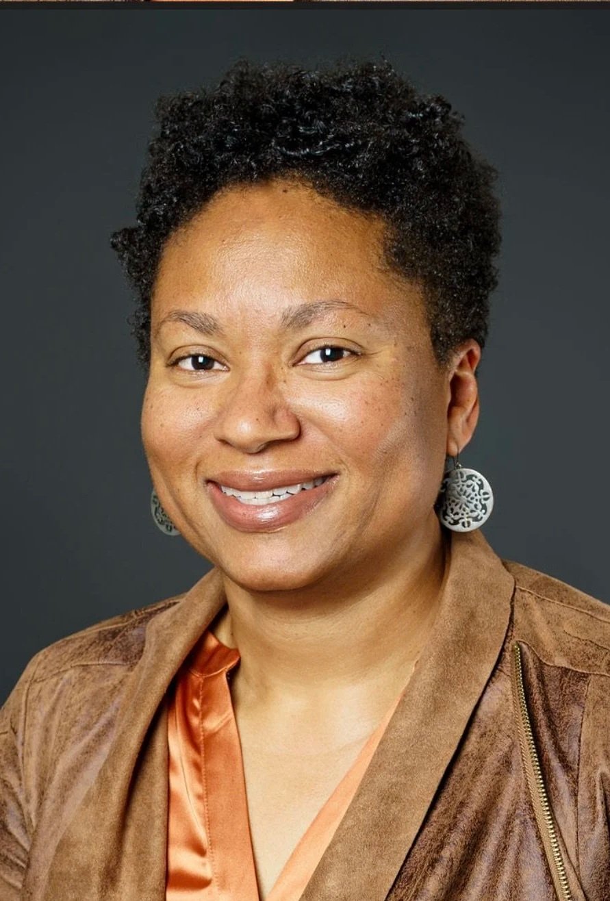 A woman with short curly black hair, wearing large circular earrings and a brown jacket over a copper-colored top, smiling against a dark background.