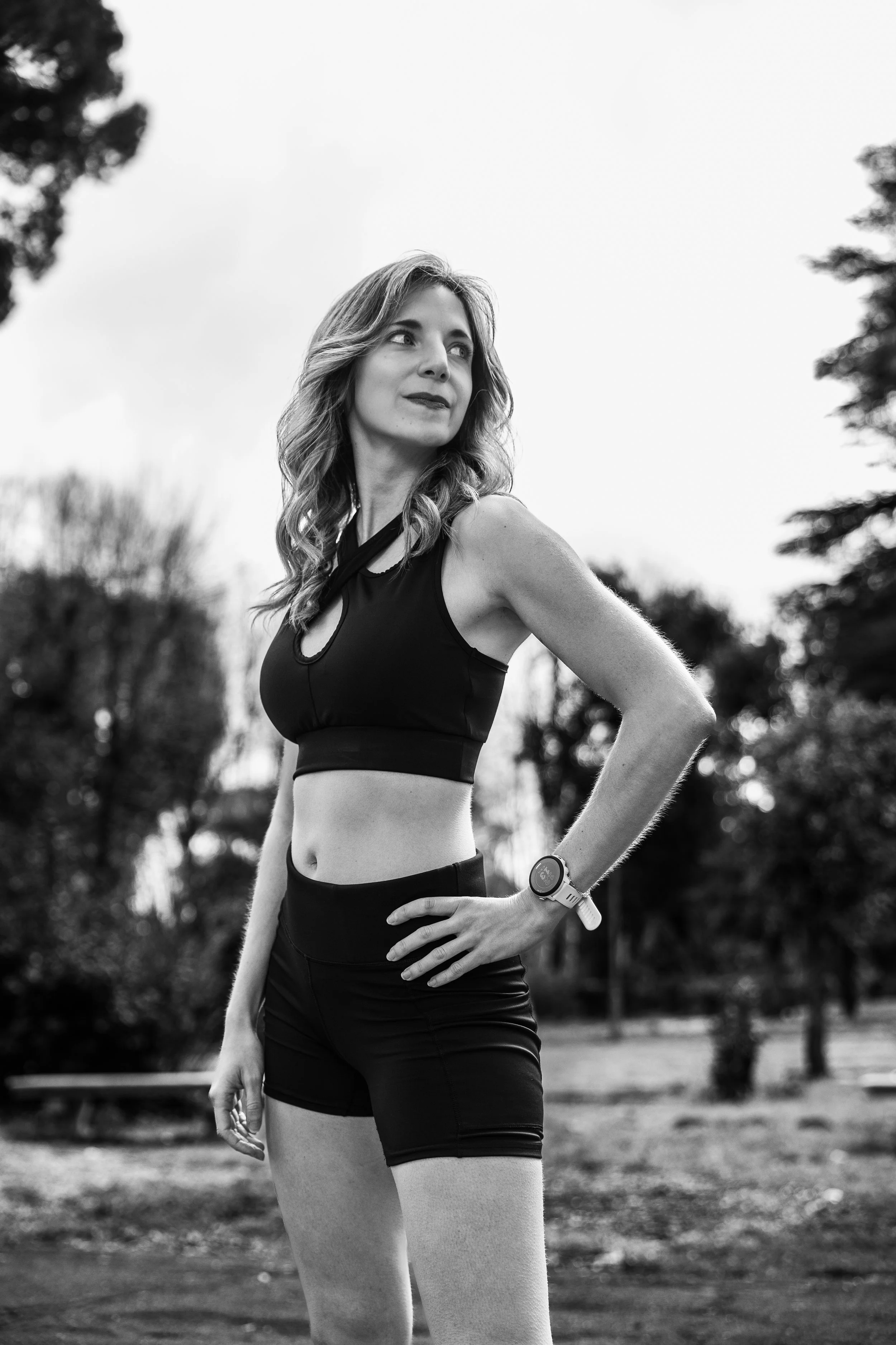A woman in athletic clothing, wearing a sports watch, standing outdoors with trees in the background, looking to her right.