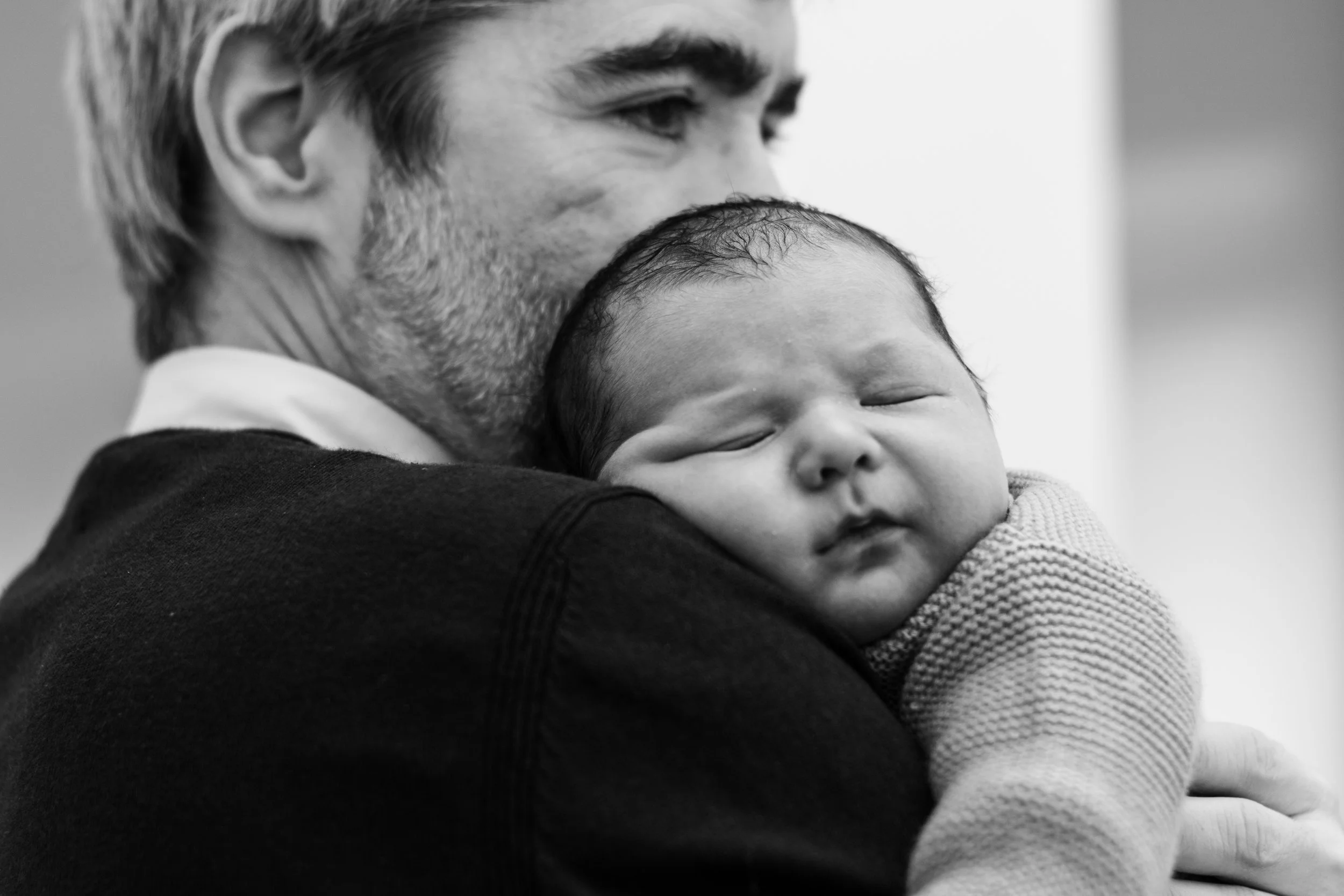 A man with gray hair and a beard, holding a sleeping baby close to his chest, with their faces touching in an intimate moment.
