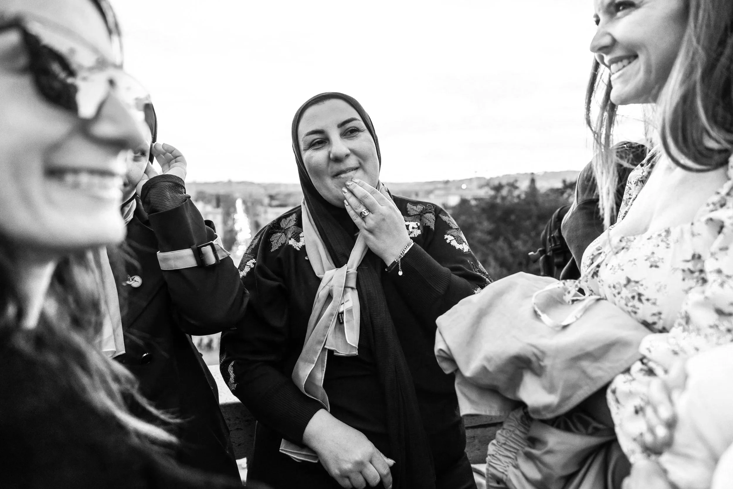 Women of diverse backgrounds are gathered outdoors, smiling and engaging in conversation, with one woman touching her chin as another holds hands in a handshake, in a candid black-and-white photo.