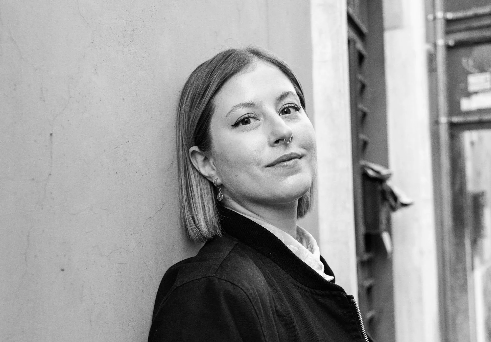 A black-and-white photo of a woman with straight hair and a septum piercing, leaning her head against a textured wall, looking softly at the camera with a slight smile.