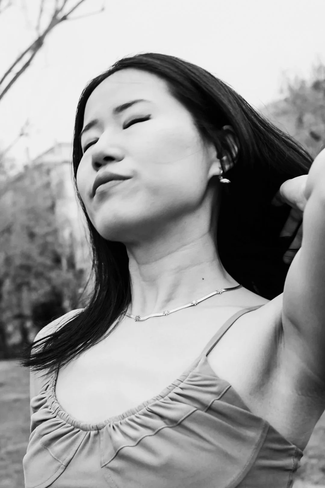 A black and white close-up photo of a woman with eyes closed, smiling slightly, holding her hair back with one hand, wearing a necklace and earrings, with trees and houses in the blurred background.