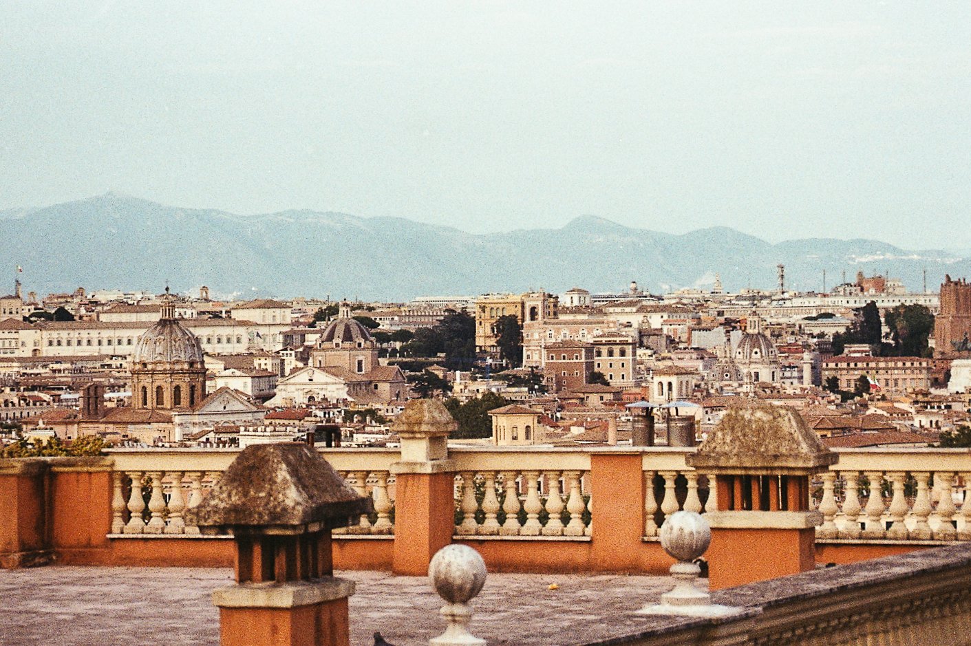 View of Rome, Italy cityscape with historic buildings and domes, mountains in the background, and a rooftop terrace with decorative balustrades in the foreground.