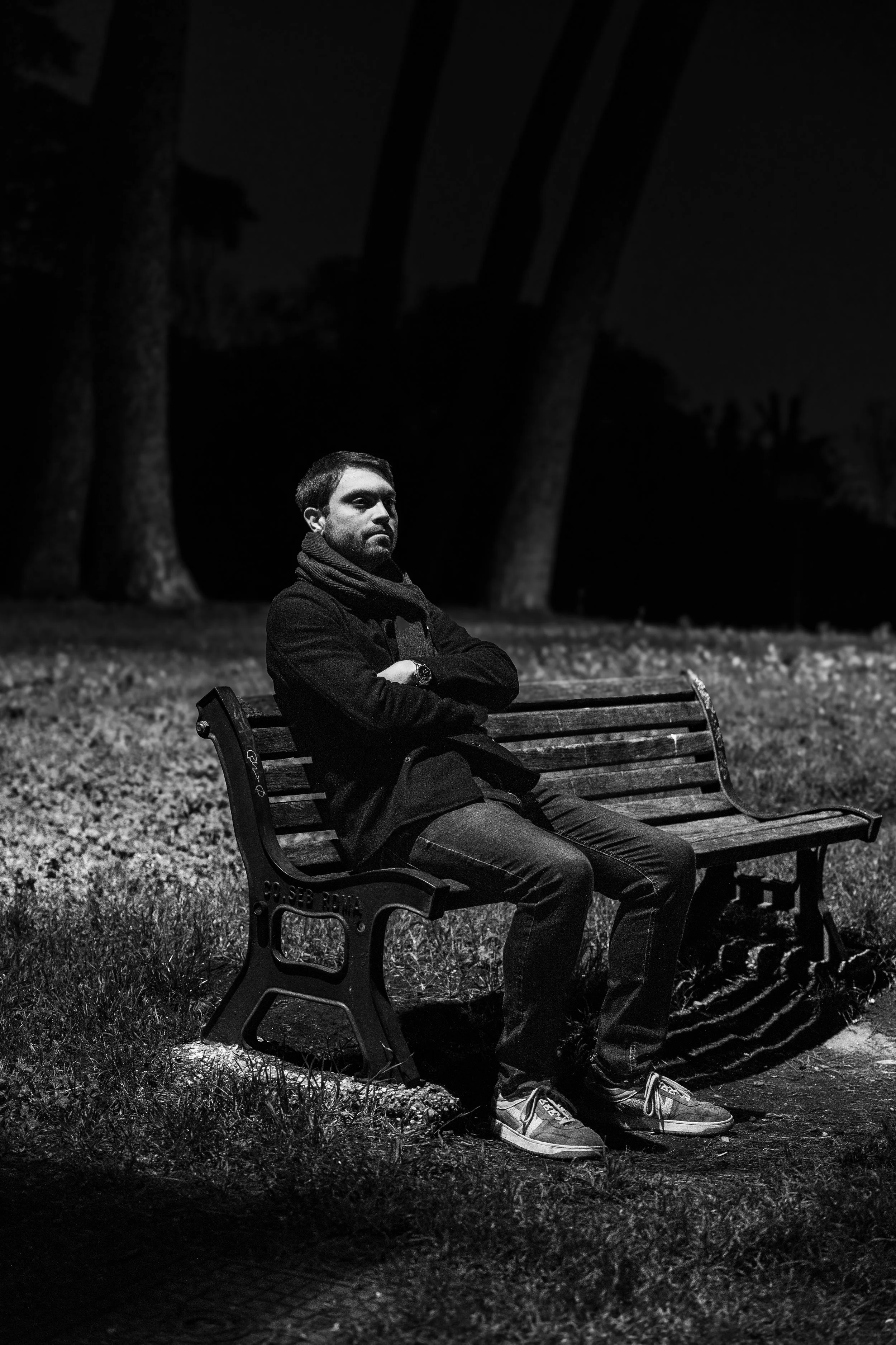 A man sitting alone on a park bench at night, with trees in the background, illuminated by a light source.