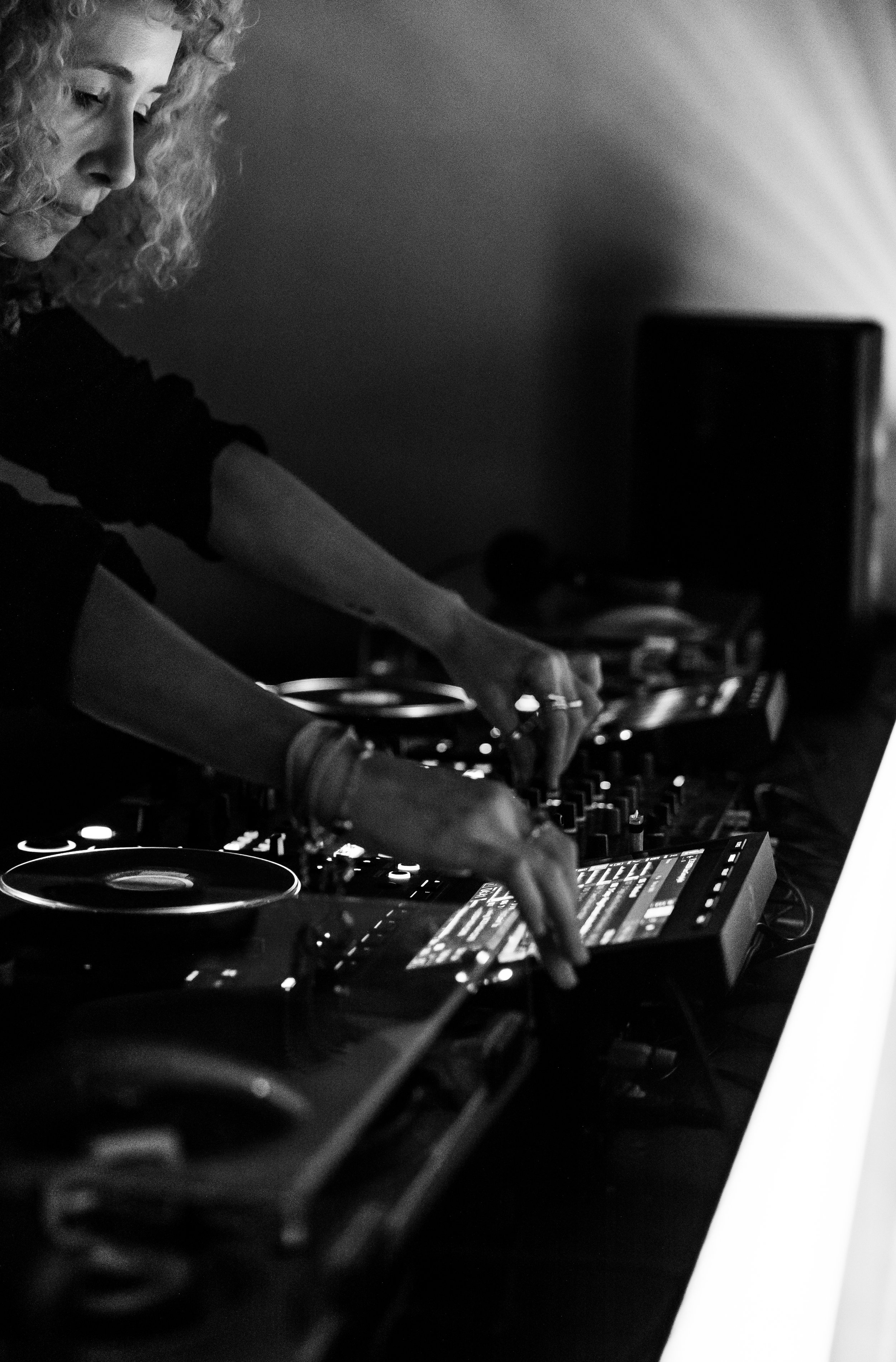A female DJ with curly hair is performing, wearing headphones and operating DJ equipment in a dark setting, with a focused expression.