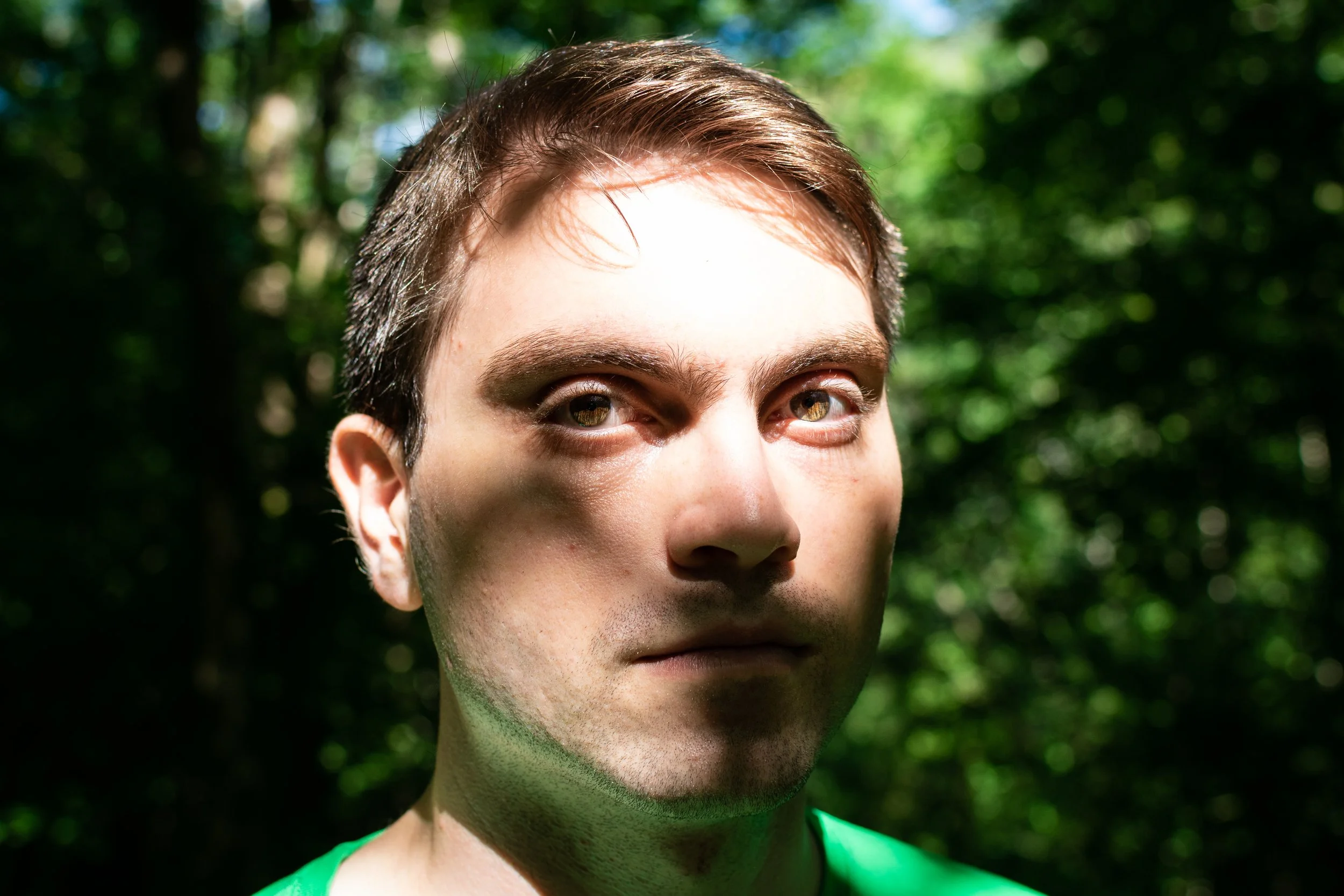 Close-up of a young man's face with brown hair and hazel eyes, outdoors with blurred green trees in the background.