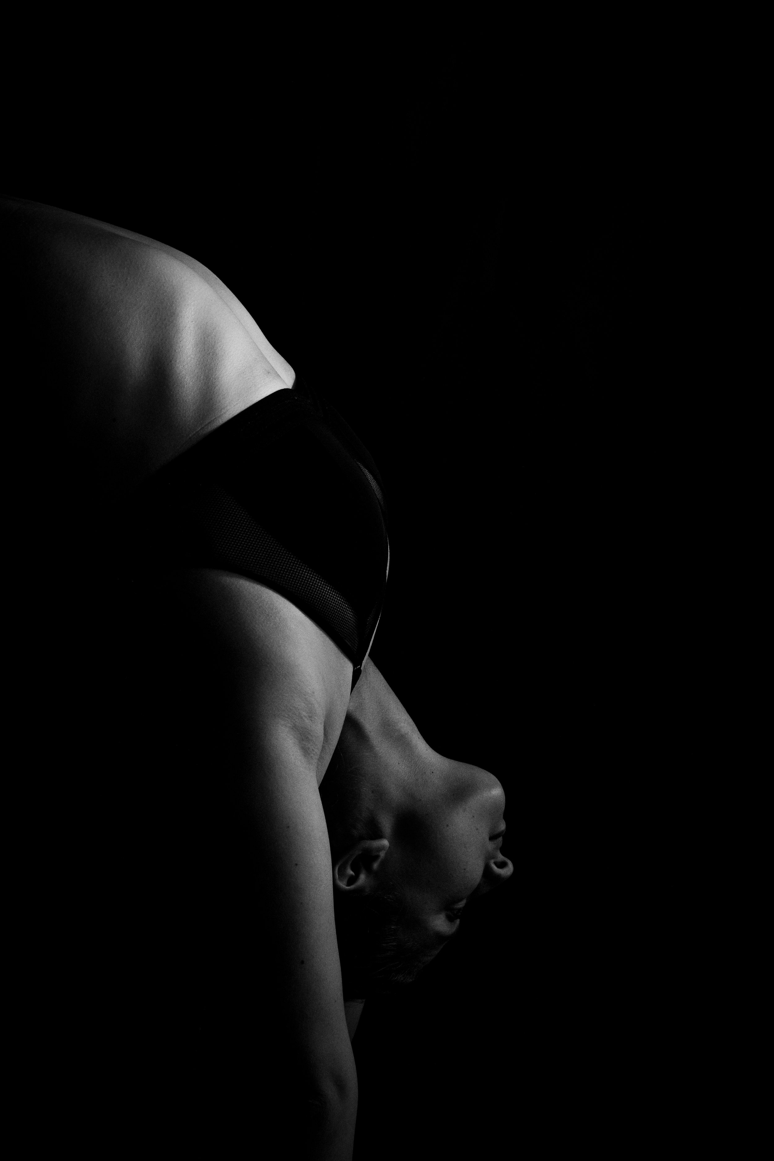 Black and white image of a woman in a yoga pose, with her head bent back, and her face turned to the side, against a dark background.