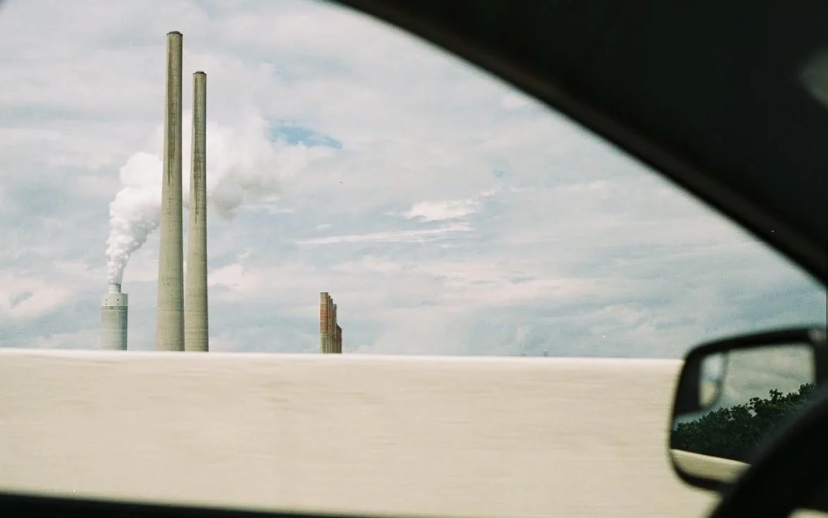 View of industrial smokestacks emitting smoke, seen from inside a car on a highway.