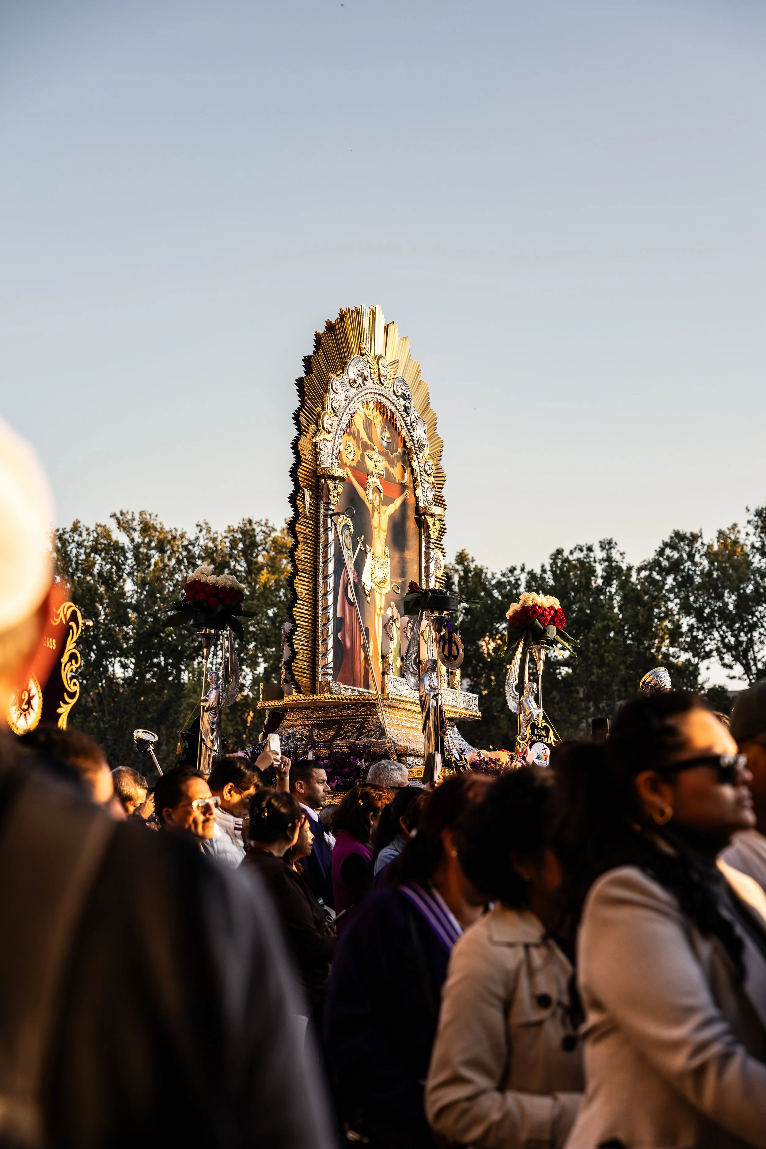 A crowd of people gathered outdoors during sunset, with a religious procession featuring a large ornate religious painting on a religious float, decorated with flowers, against a backdrop of trees and a clear sky.