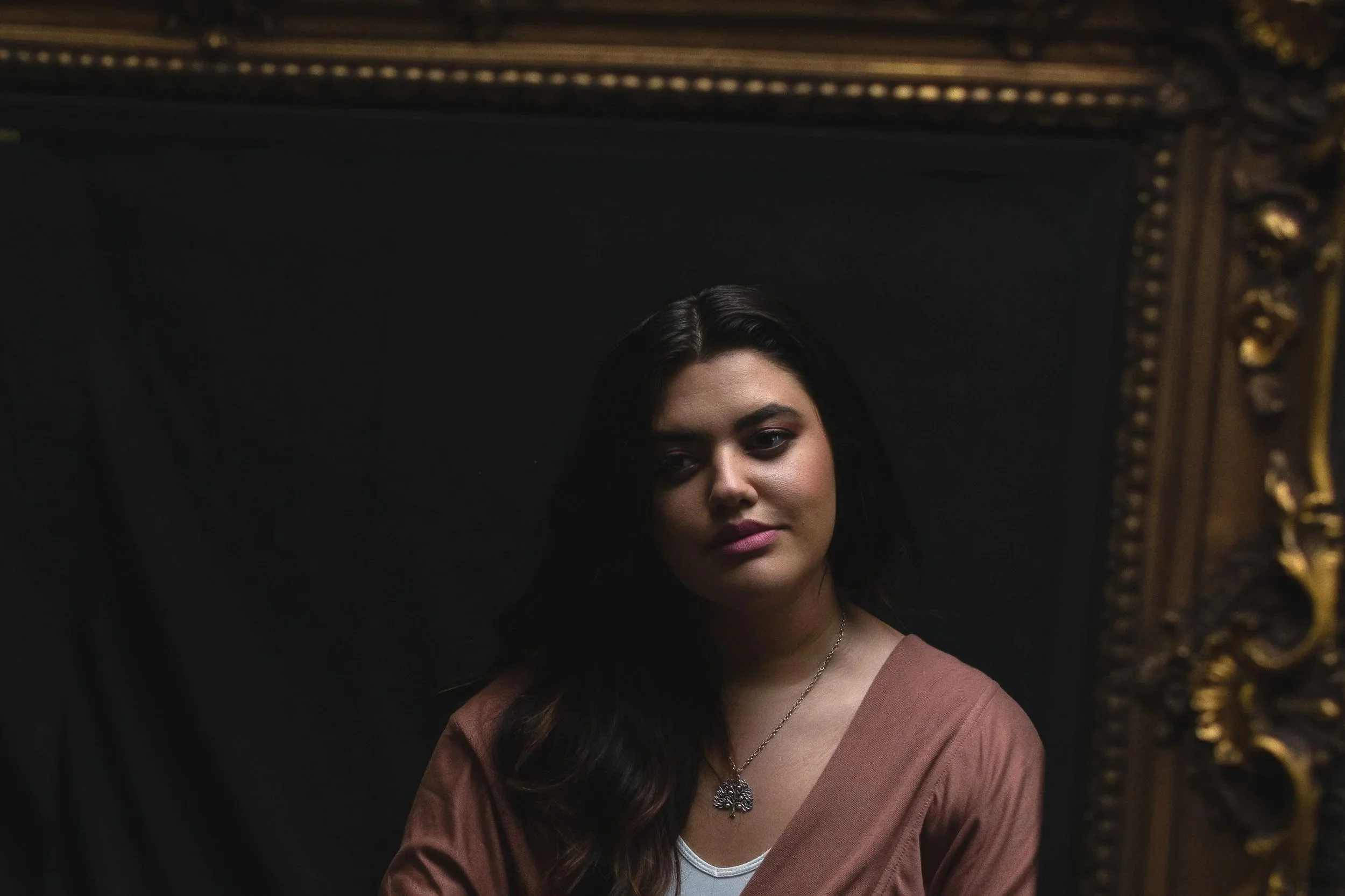 A woman with long dark hair and light skin wearing a brown top and a pendant necklace, standing in front of a dark background with a gold ornate frame or decoration on the right side.