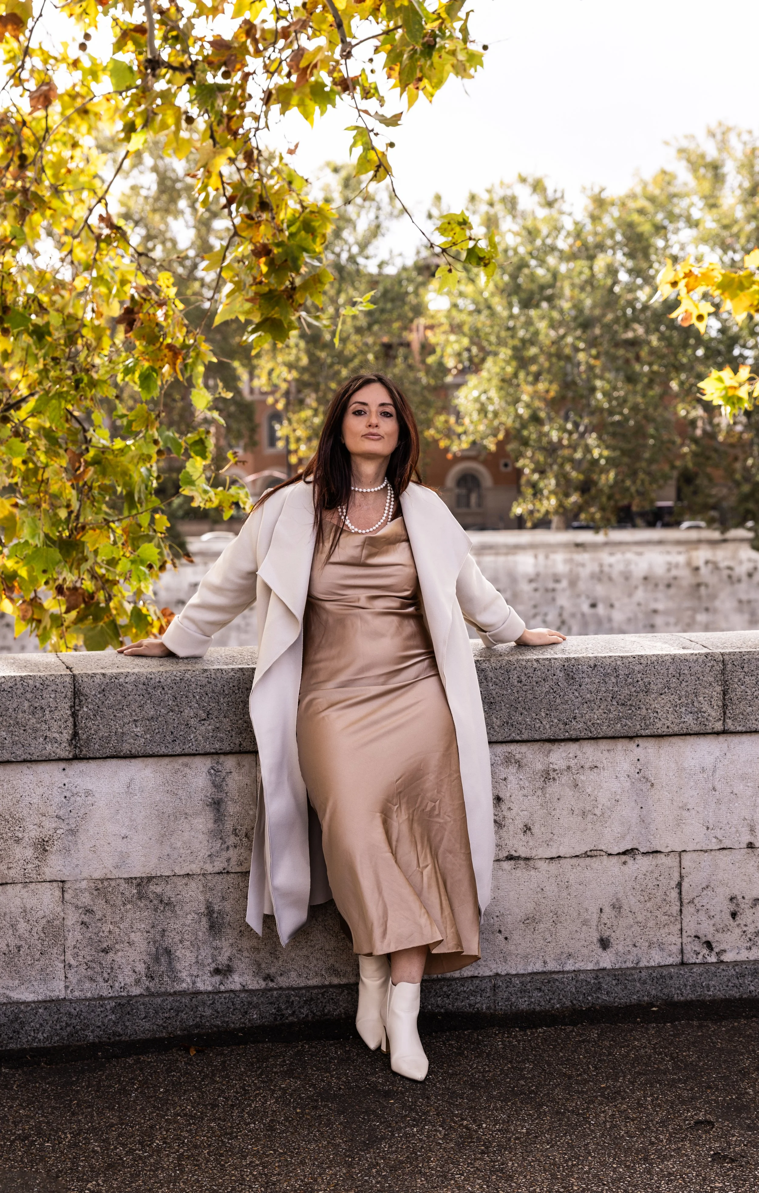 A woman wearing a beige dress, cream blazer, pearl necklace, and cream ankle boots standing against a stone barrier with autumn leaves in the background.