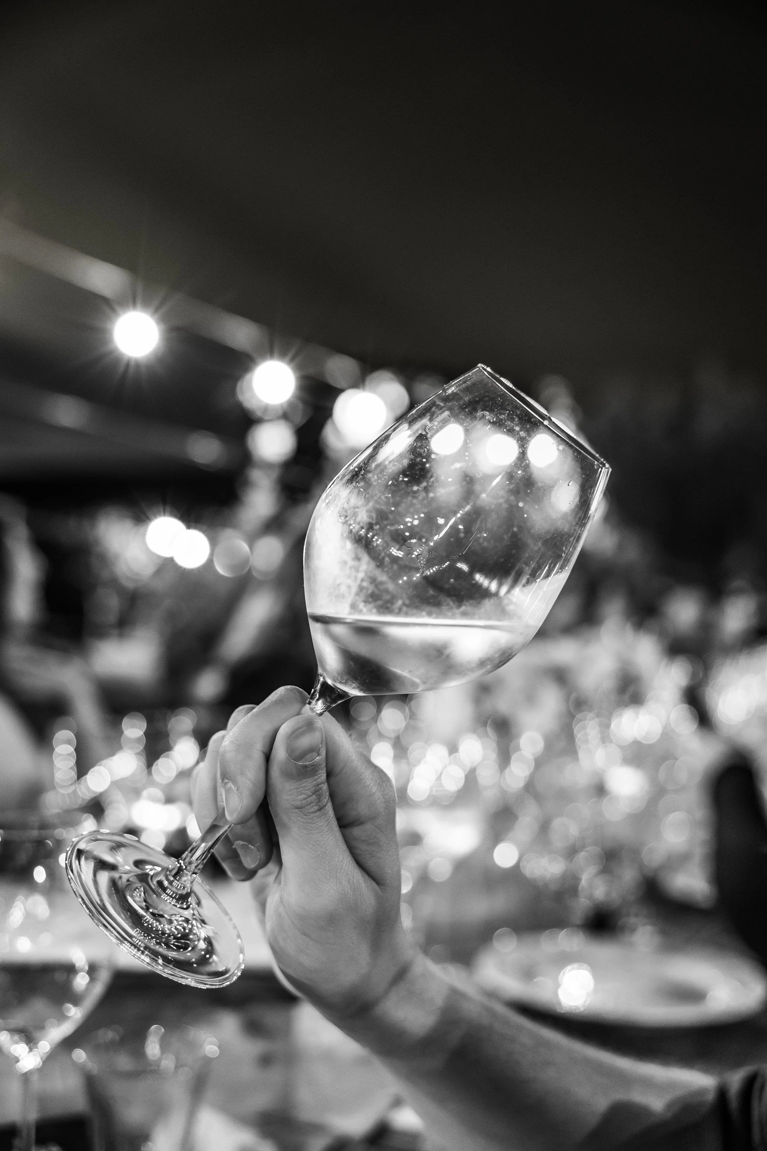 A hand holding a wine glass filled with a clear beverage, with blurred lights in the background.