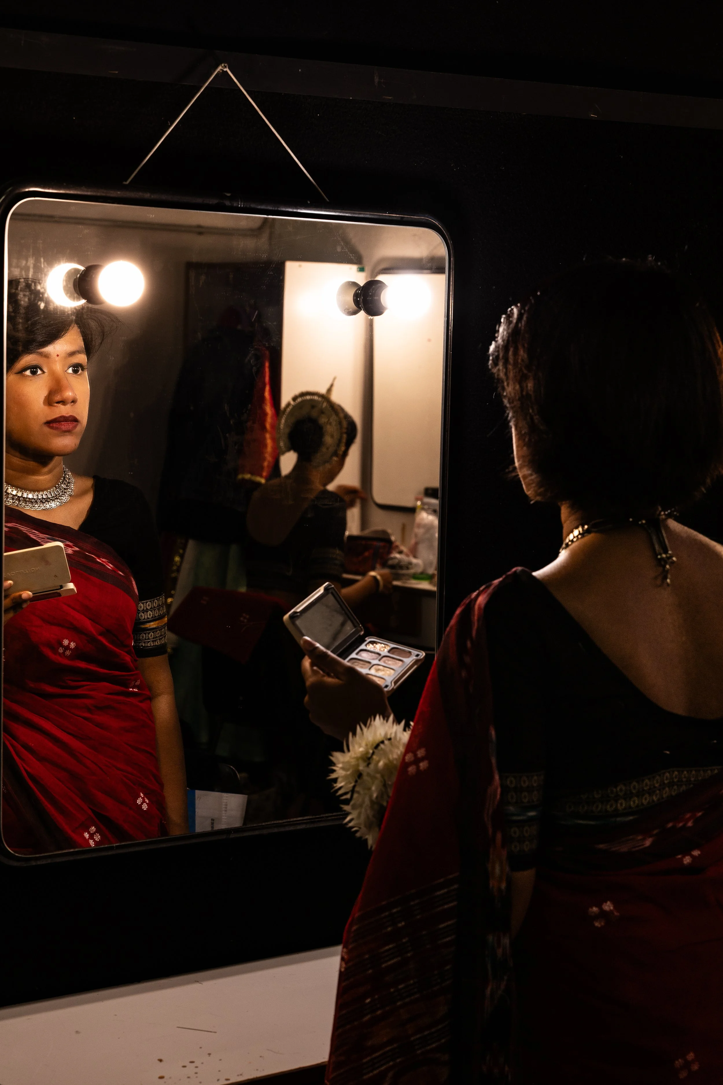 A woman dressed in traditional red and black attire looking into a mirror with lights, holding a makeup palette, with her reflection visible in the mirror.