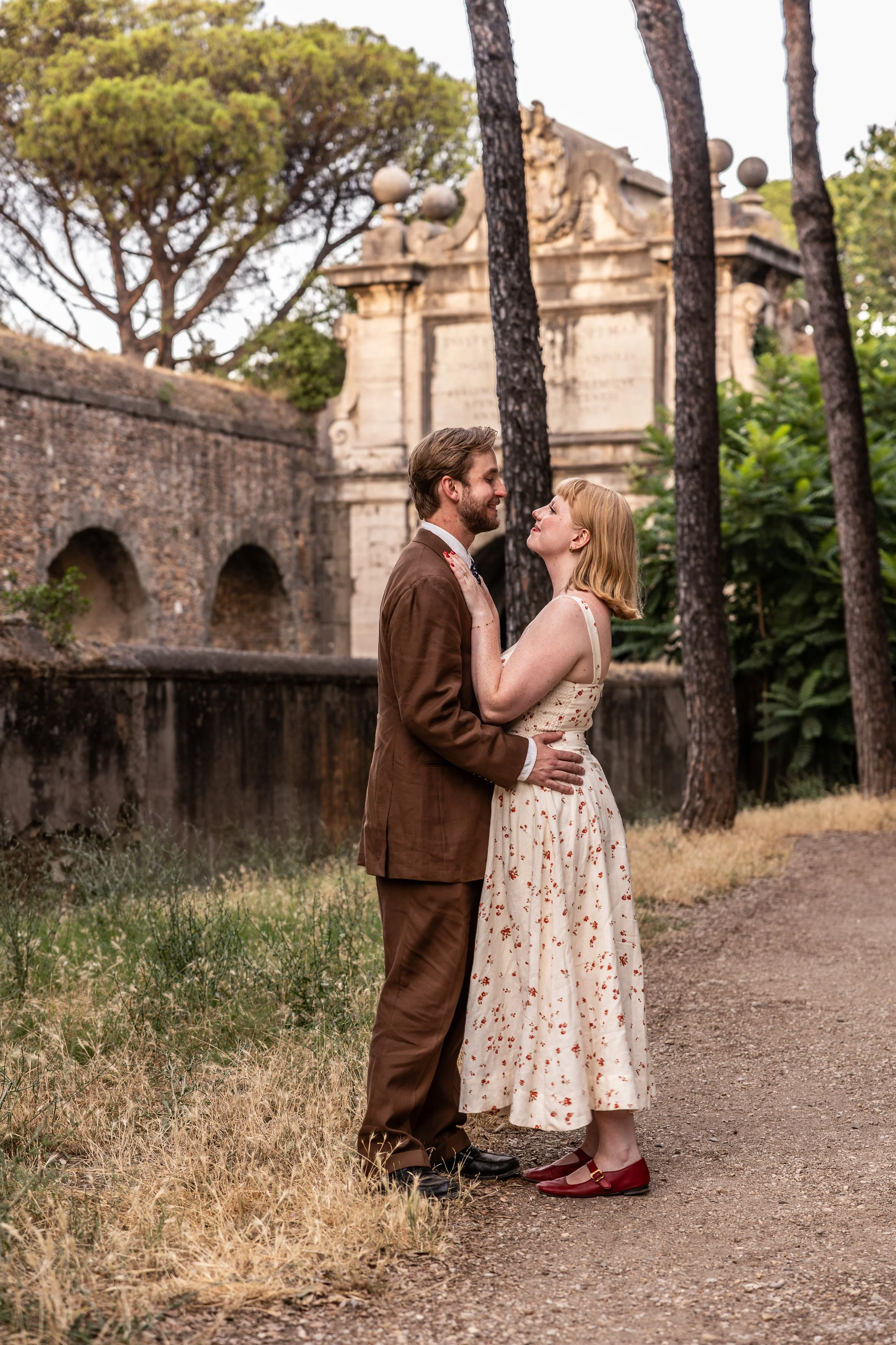 A man and woman stand close together outdoors, smiling and gazing into each other's eyes. The man wears a brown suit and the woman wears a vintage floral dress with red shoes. They are in front of trees, a stone wall, and an old monument or building 