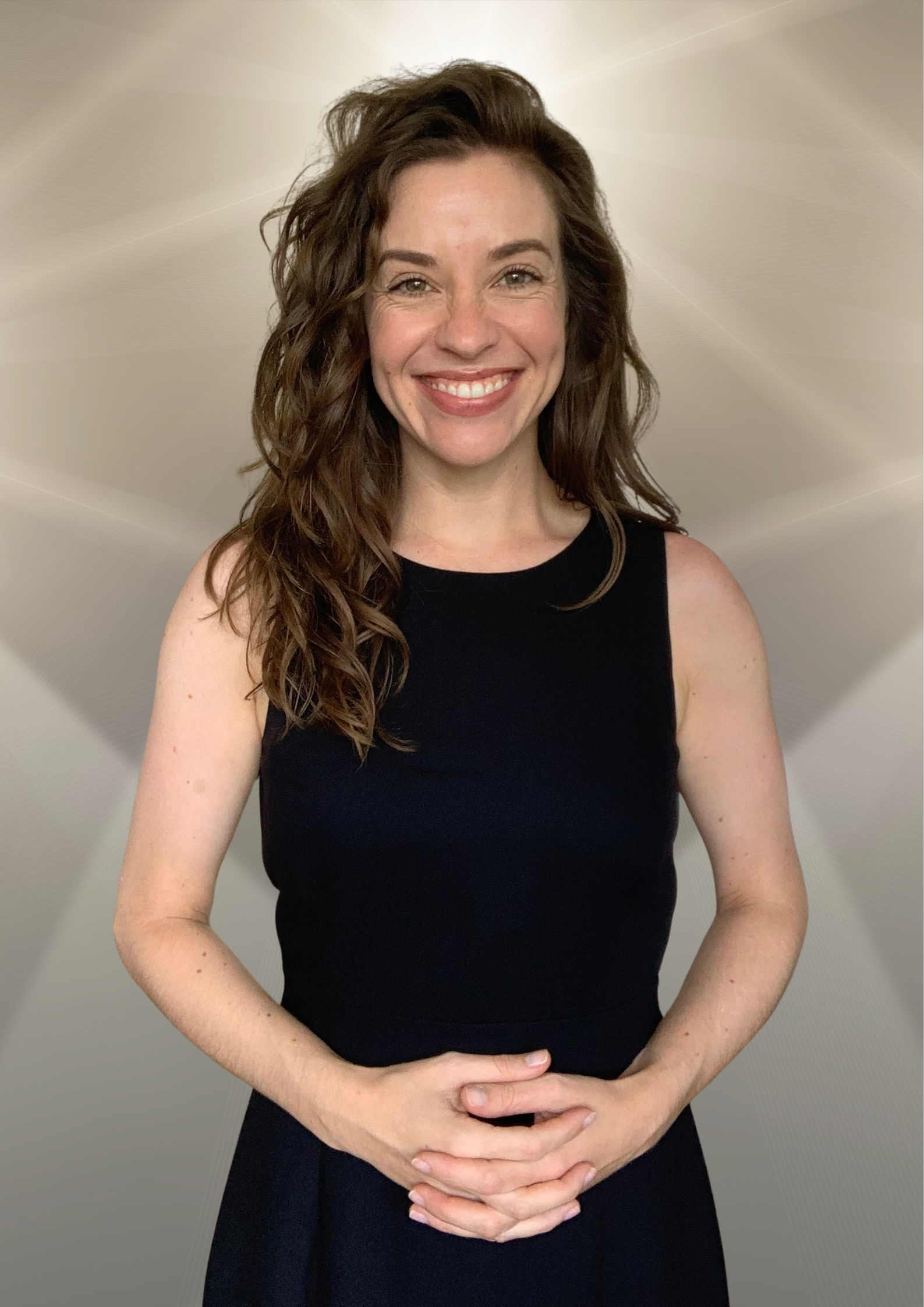 A woman with curly brown hair wearing a sleeveless black top, standing against a neutral patterned background, smiling with her hands clasped in front of her.