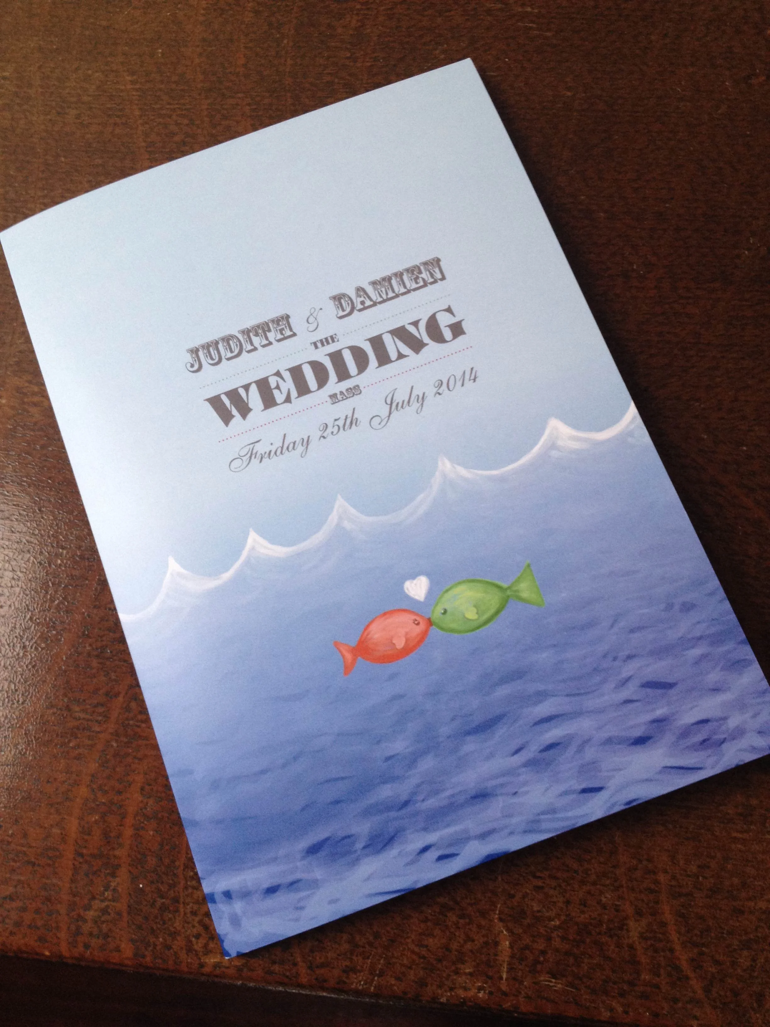 A wedding invitation featuring a blue ocean background with two cartoon fish, one orange and one green, facing each other with a small heart between them. The text includes 'Judith & Damien,' 'The Wedding,' 'Friday 25th July 2014.'
