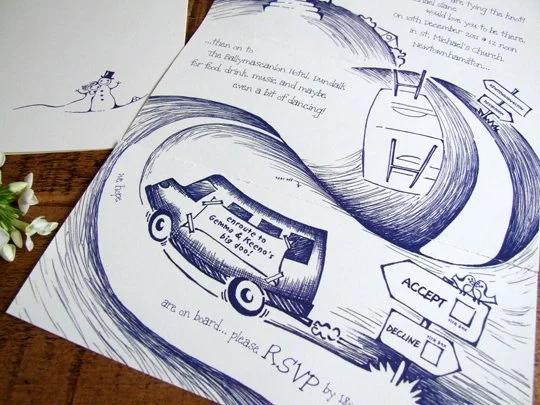A printed wedding invitation featuring a cartoon-like illustration of a bus with mountains in the background, and a winding road. The invitation includes RSVP details, an acceptance box, a decline box, and decorative elements.