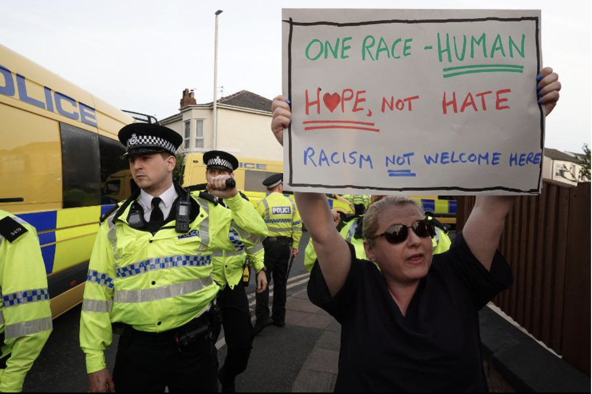A white woman holding a sign that says "One race - Human, Hope not hate, Racism not welcome here", surrounded by police during a protest against far-right violence