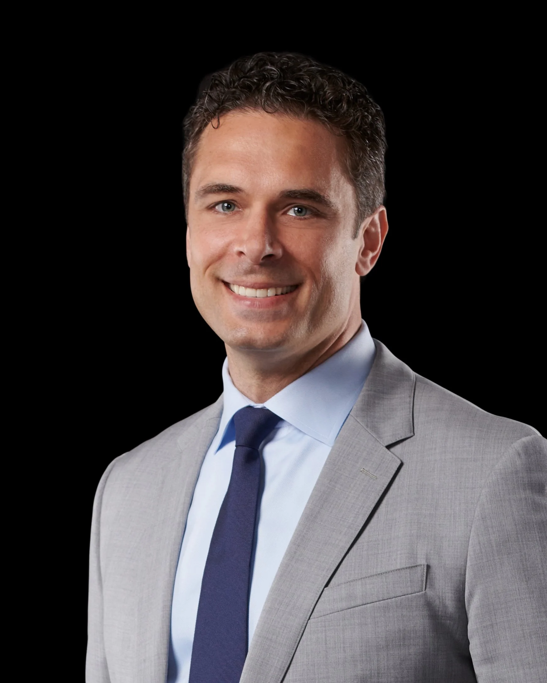 Headshot of a smiling Raffaele Villella with curly brown hair, wearing a light gray suit with a white shirt and navy blue tie, against a black background.
