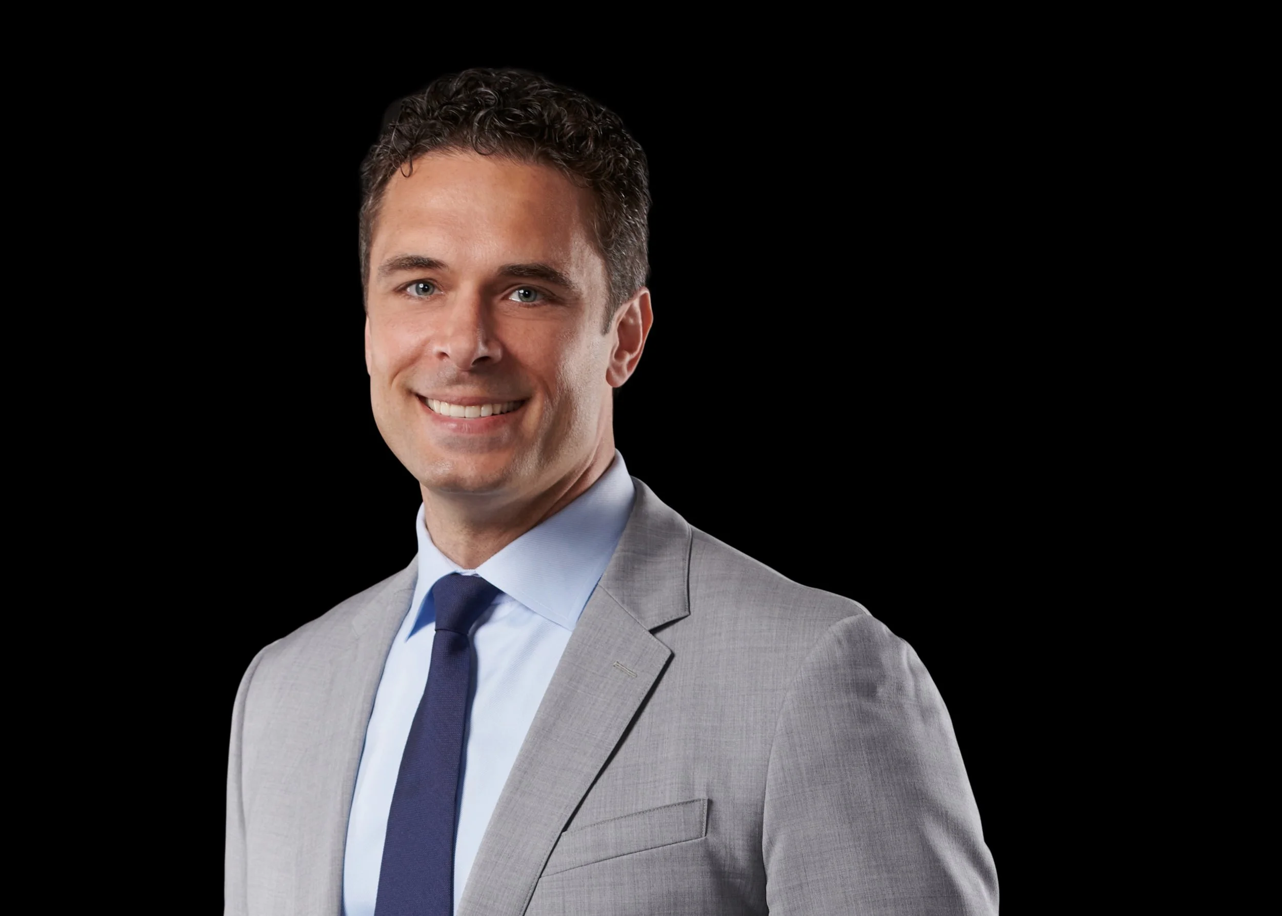 Portrait of a smiling Raffaele Villella in a gray suit with a blue tie, against a black background.