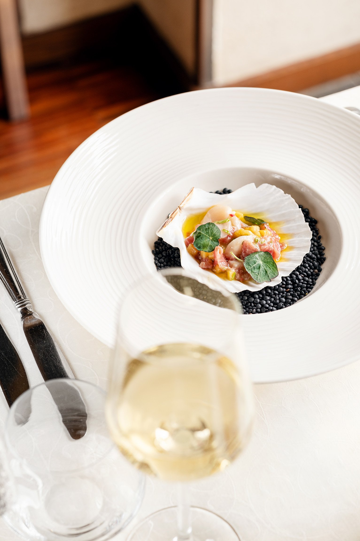 A white plate with black lentils and a shell with a seafood mixture, garnished with green herbs, accompanied by a glass of white wine.