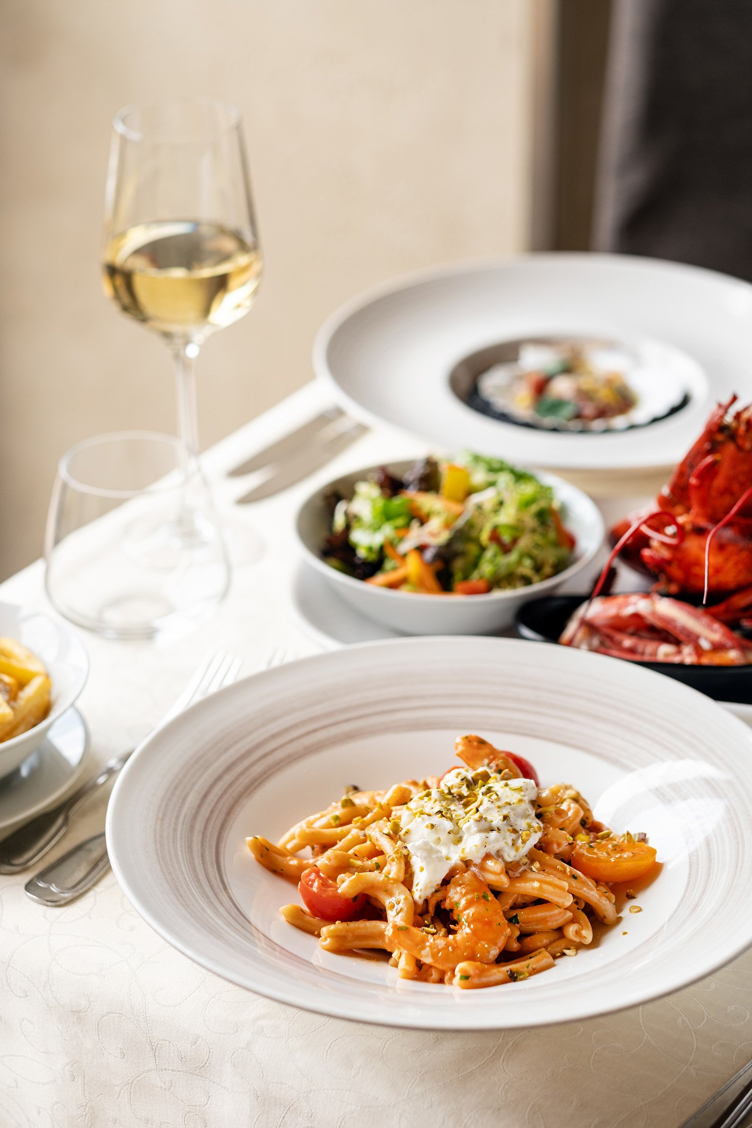 A table set with various dishes including a plate of pasta with shrimp and cherry tomatoes, a salad, and a seafood platter, with a glass of white wine.