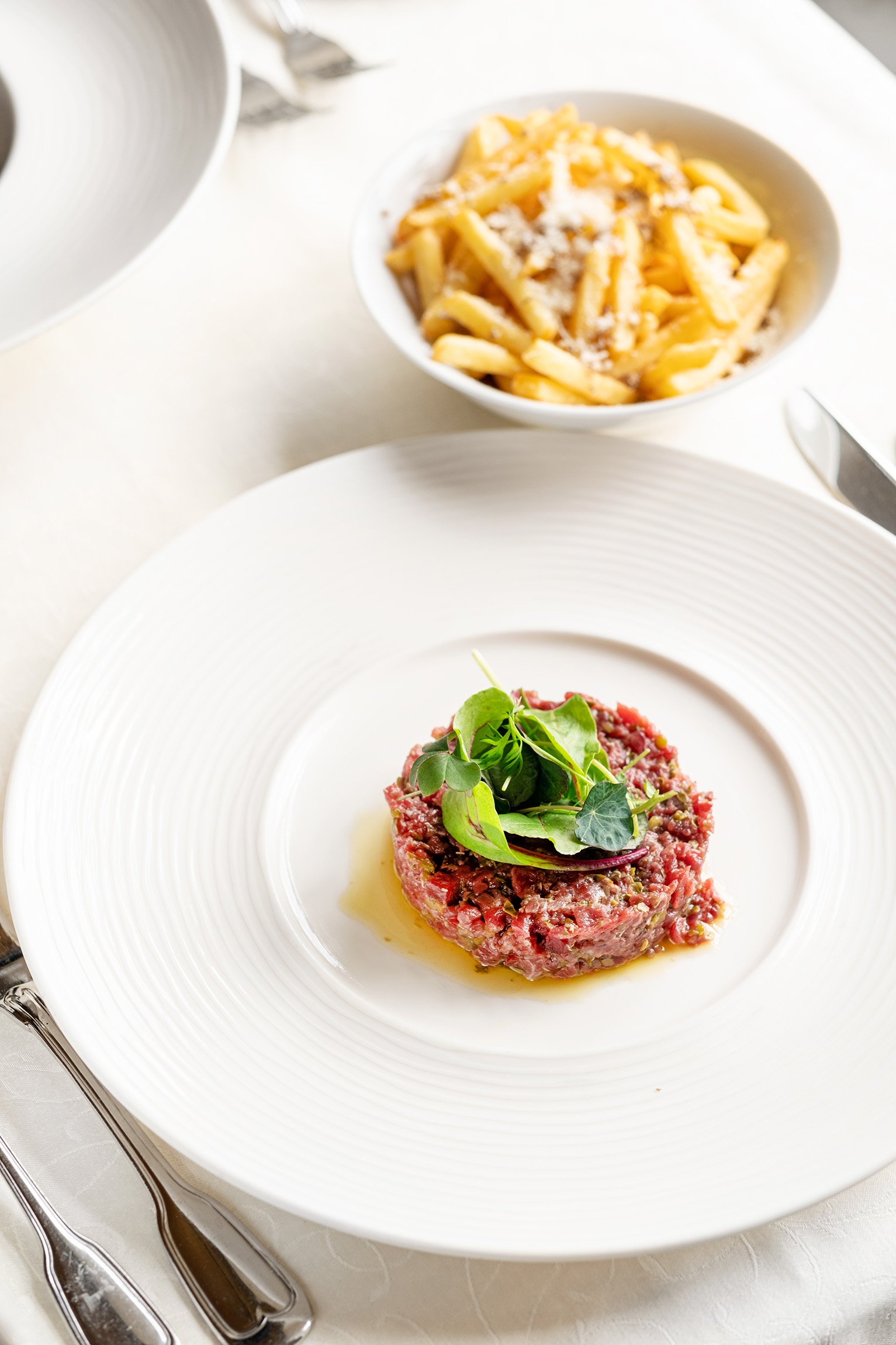 A plate of steak tartare garnished with microgreens and a bowl of pasta topped with grated cheese in the background.