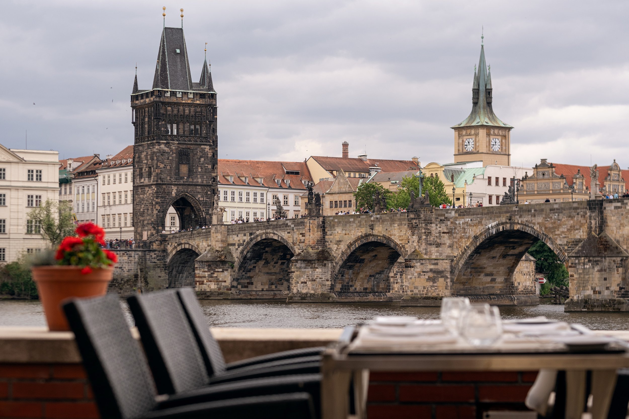 View of Charles Bridge and Old Town Bridge Tower in Prague, Czech Republic, with tables in the foreground at a riverside cafe, overcast sky.