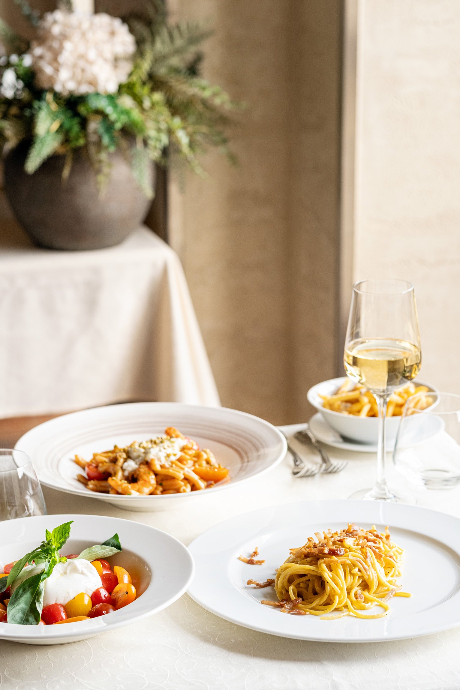 A table with two plates of pasta, a salad with cherry tomatoes and basil, a glass of white wine, and a bowl of fries, set in a well-lit room with a flower arrangement in the background.