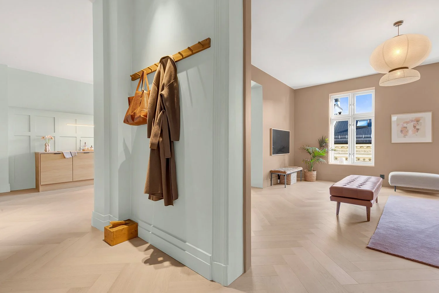 Interior view of a modern living room with large windows, beige walls, and wooden flooring, featuring a pink ottoman, a white bench, and a potted plant.