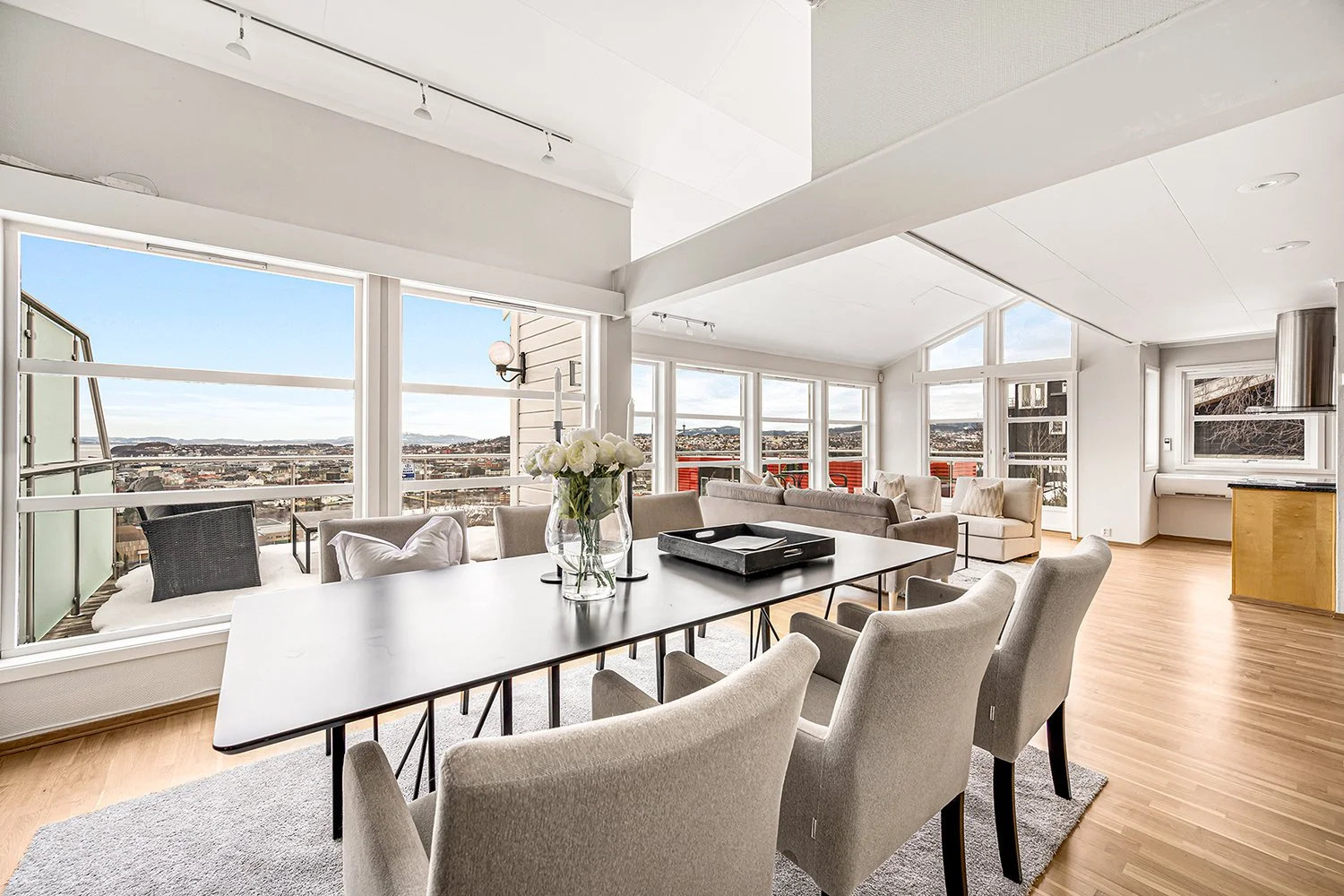 Open-plan living and dining area with large windows overlooking cityscape, featuring a dining table with six upholstered chairs, a vase with white flowers, a gray sofa, and additional seating near balconies with red railings.