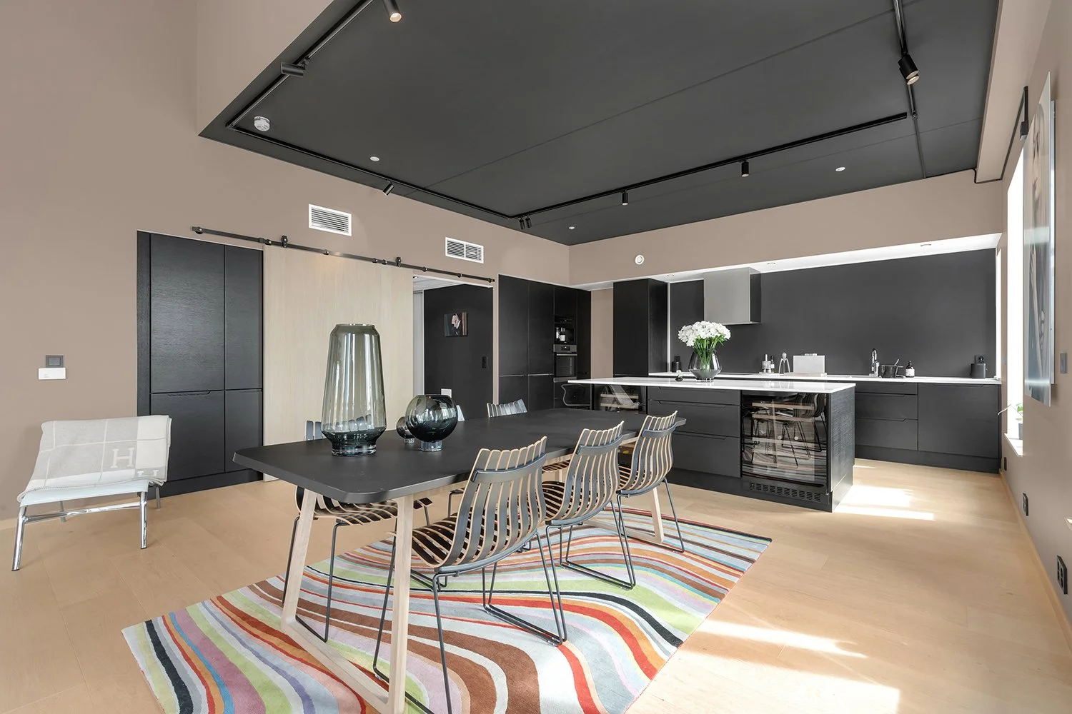 Modern kitchen and dining area with black cabinetry, white countertops, a black table with wooden chairs, a colorful striped rug, and decorative vases on the table.