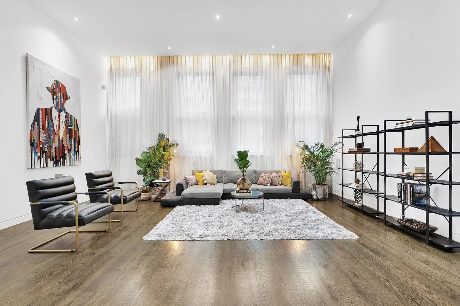 Modern living room with white walls, large windows with sheer curtains, and dark hardwood floors. Features a light gray sectional sofa with colorful pillows, a round coffee table with a plant, and two black and gold modern chairs. There is a white sh