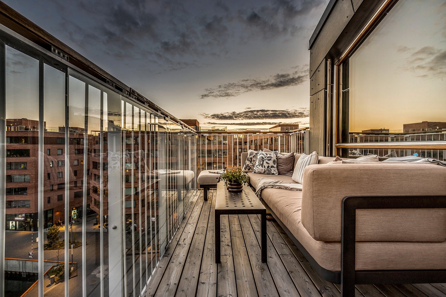 A balcony with a beige sofa, pillows, and a coffee table with a potted plant, overlooking a cityscape at sunset.