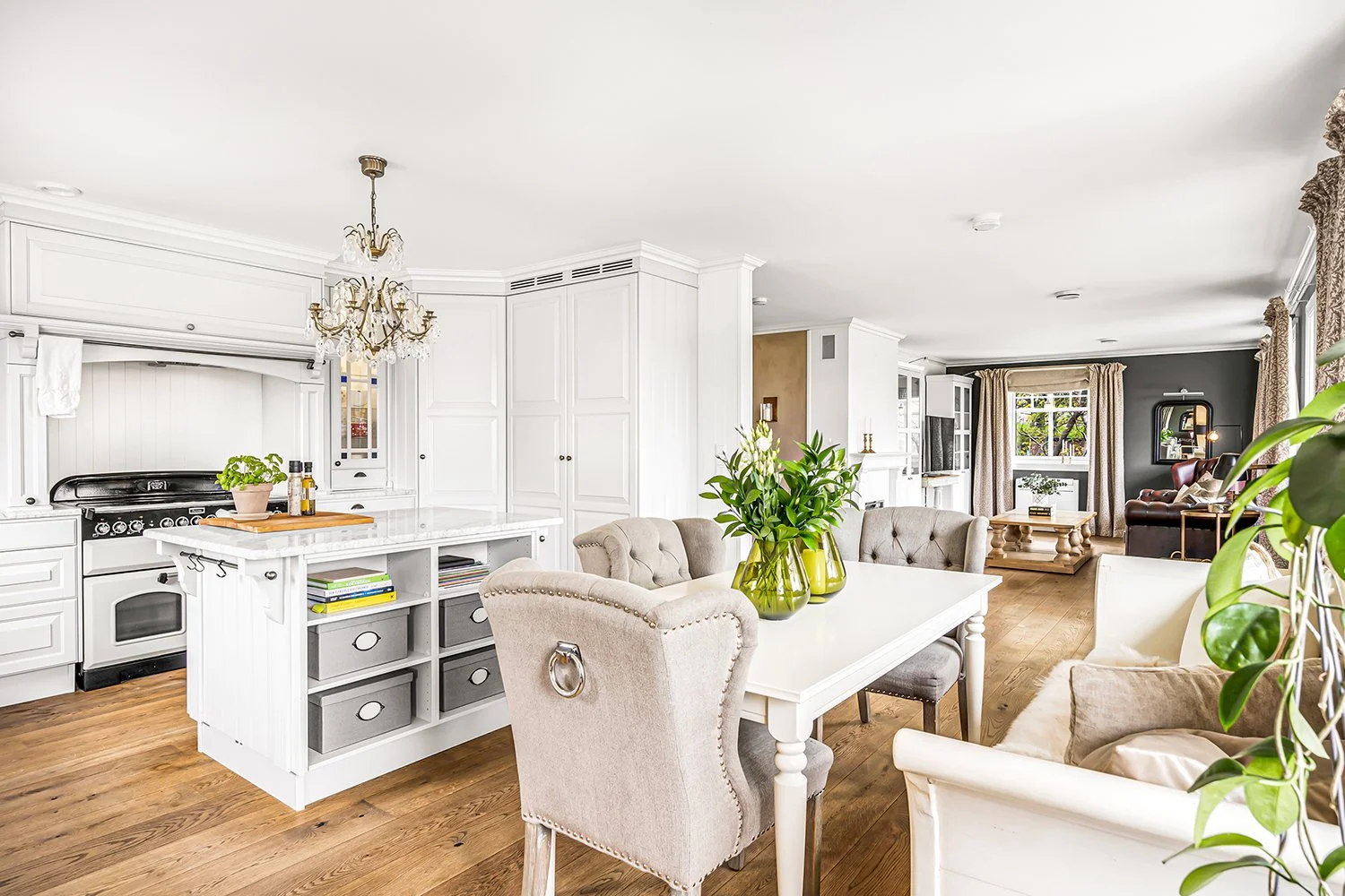 Open-concept living and dining room with white and beige furniture, wooden flooring, green plants, and large windows with curtains, in a bright, modern home.
