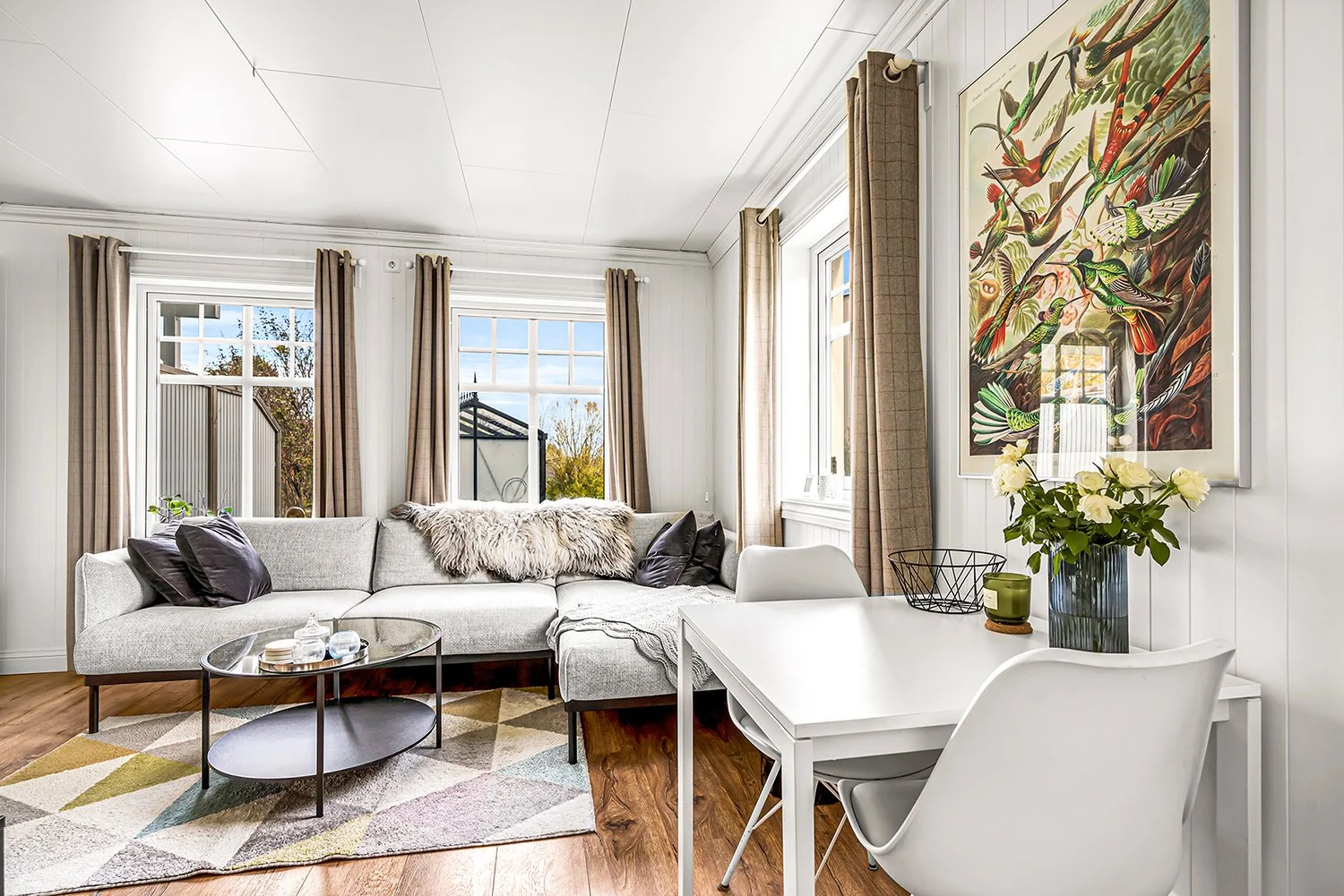 Living room with a gray sectional sofa, a multicolored rug, a black oval coffee table, white paneled walls, large windows with beige curtains, a white dining table with white chairs, a vase with white flowers, wall art, and hardwood flooring.