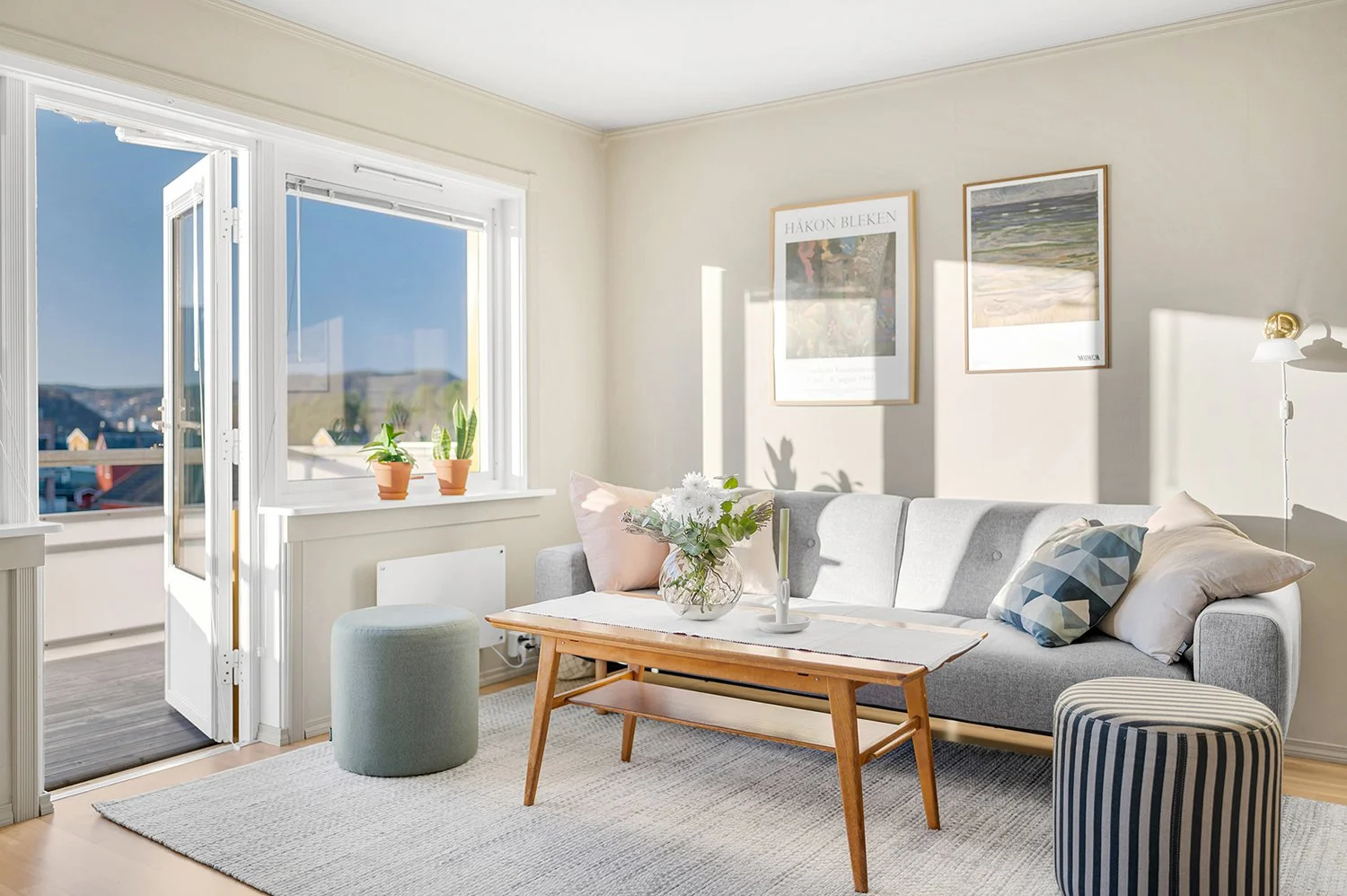 Bright living room with open balcony door, sunlight streaming in, gray sofa with pillows, wooden coffee table with flowers, two round ottomans, framed artwork on wall.