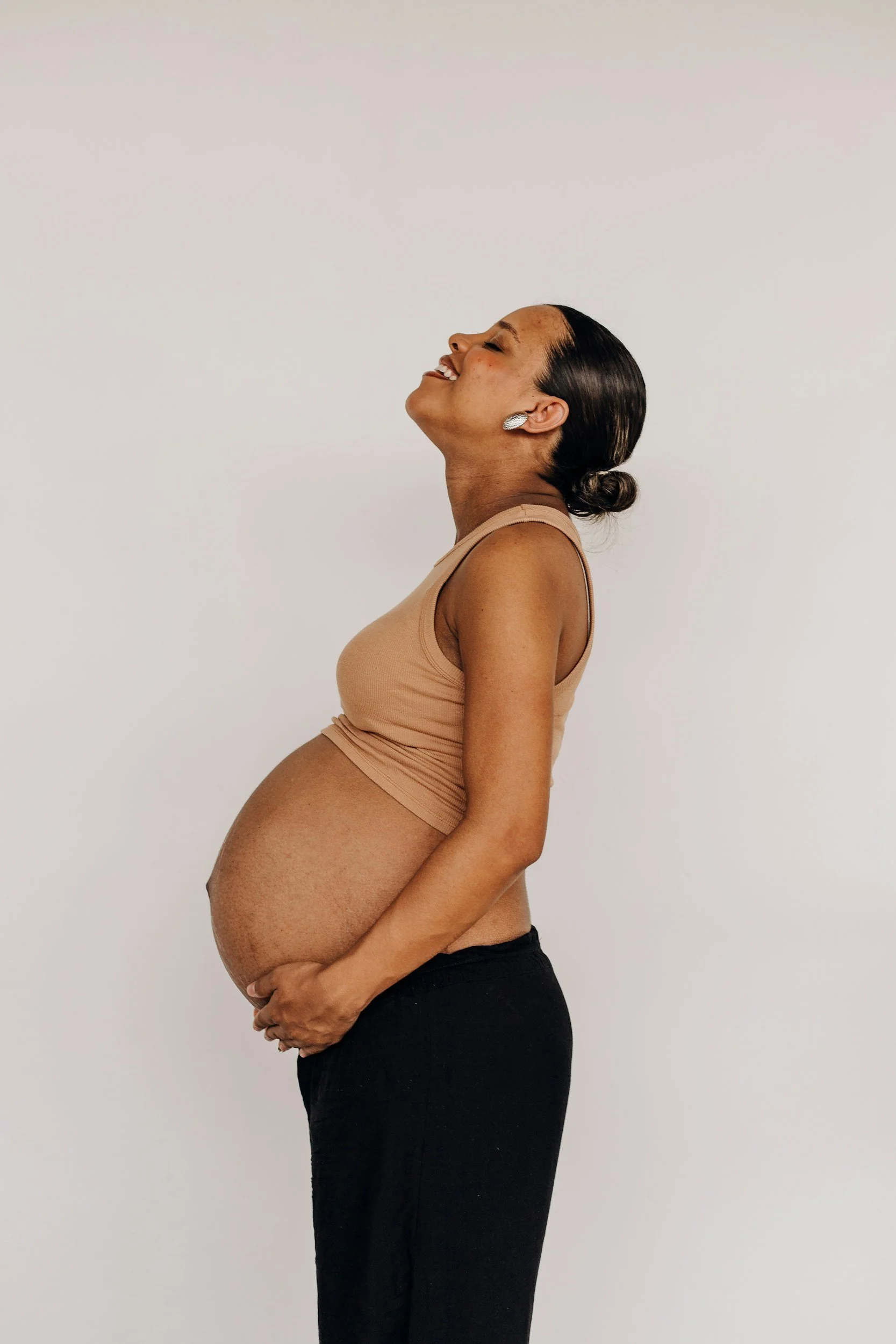 Pregnant woman smiling with eyes closed, wearing a beige sleeveless top and black pants, holding her belly against a plain white background.