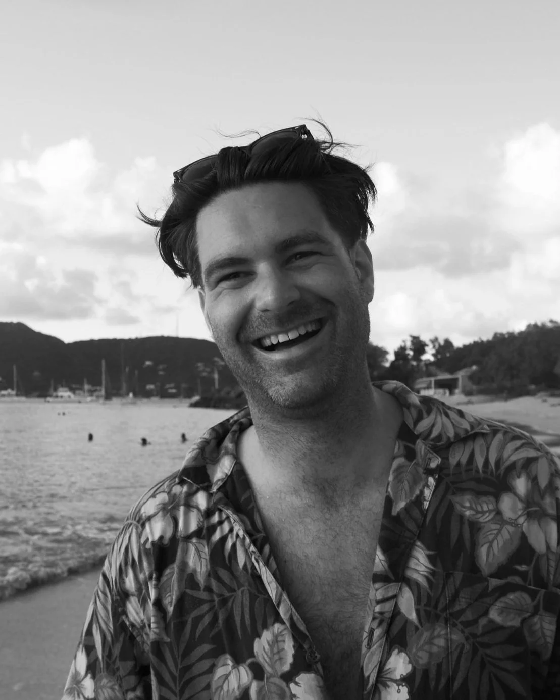 A smiling man with sunglasses on his head wearing a Hawaiian shirt, standing at the beach.