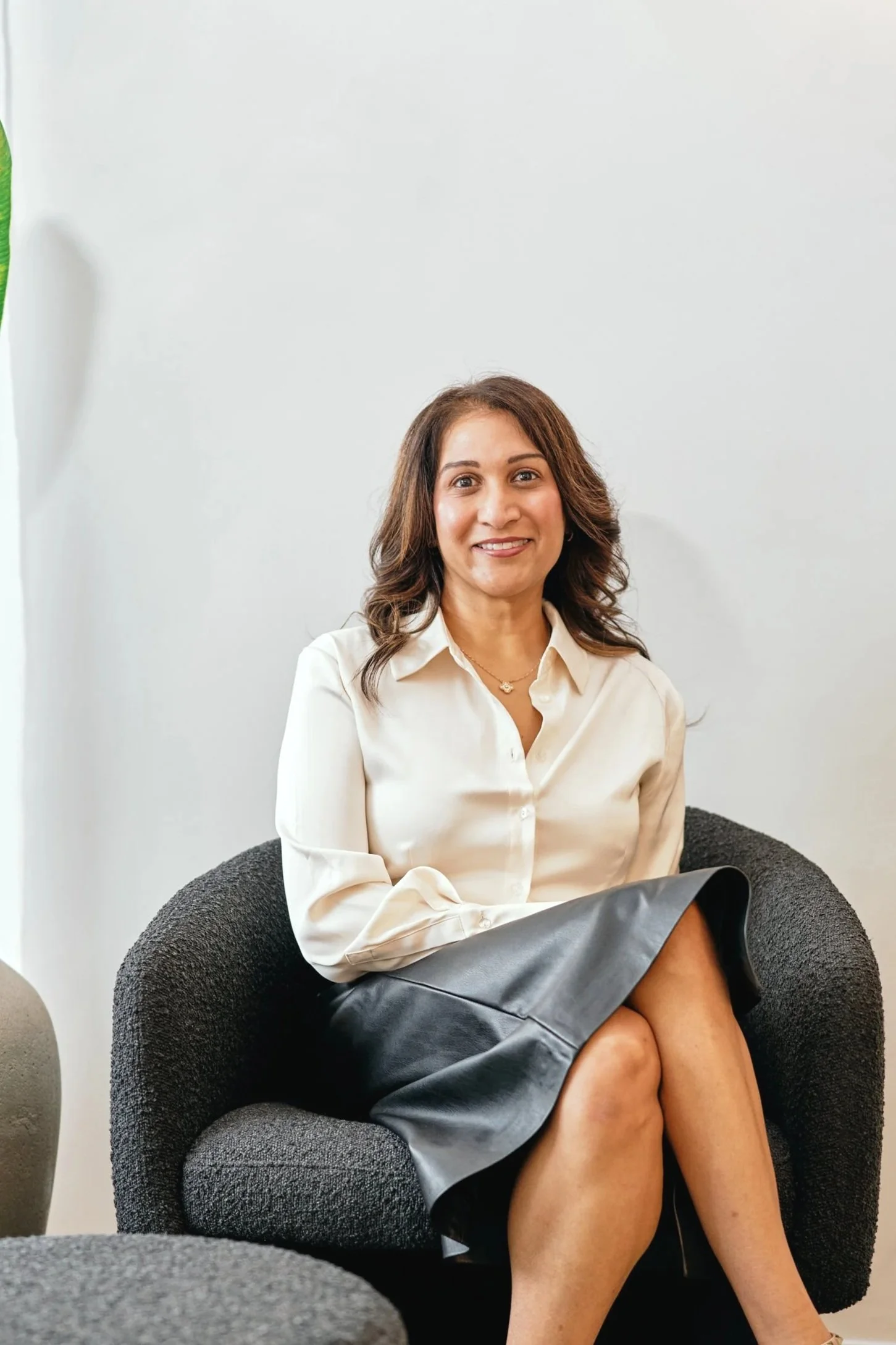 A woman with shoulder-length brown hair, wearing a white blouse and a gray skirt, sitting on a dark gray textured armchair with her legs crossed. She is smiling and looking at the camera, in a bright sitting area.