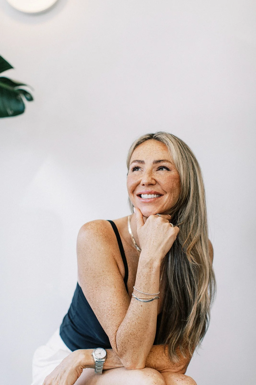 A woman with blonde hair, freckles, and a bright smile, wearing a black tank top and jewelry, sitting and looking up in a white room.