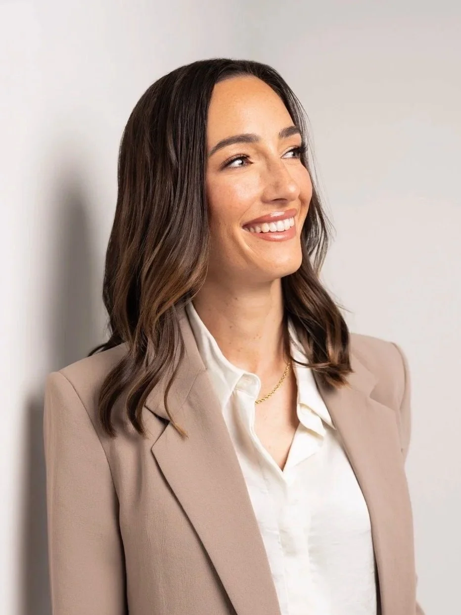 A woman with medium-length brown hair styled in loose waves, wearing a beige blazer, white blouse, and a gold necklace, smiling while looking to her left.