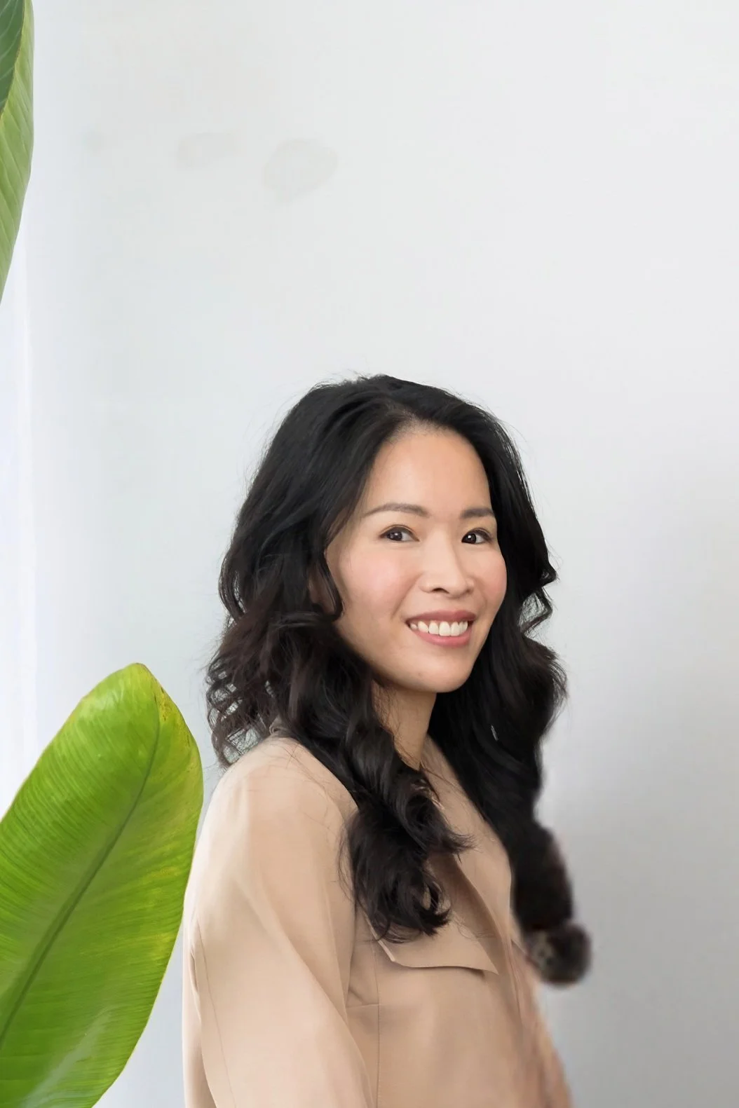 A woman with long, black, curly hair smiling against a plain white wall, with a large green leaf partly visible in the foreground.