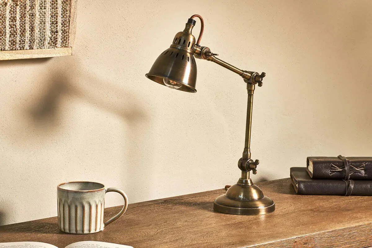 A brass adjustable desk lamp, a ceramic mug, and black books on a wooden desk against a plain wall.